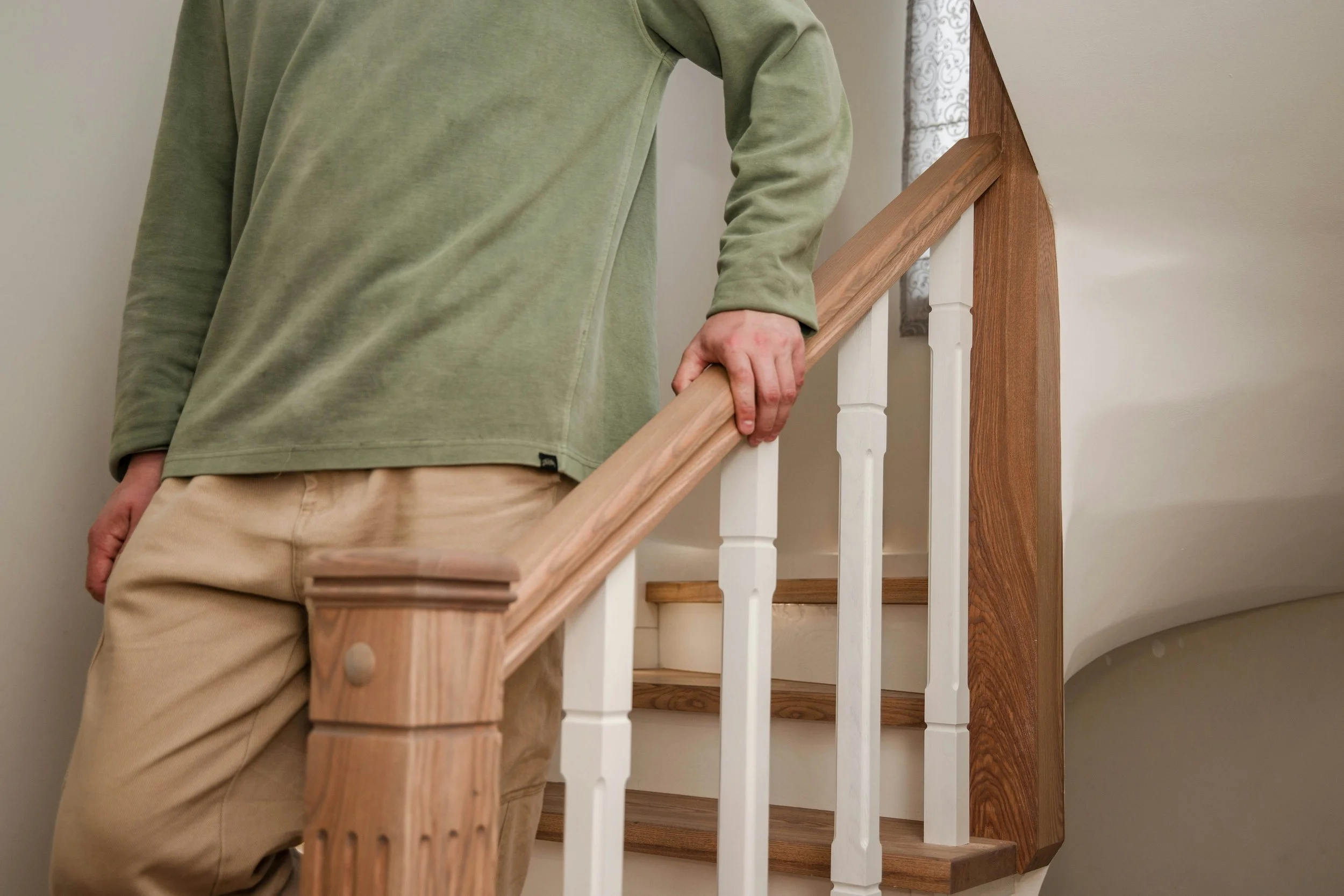 A person holding a wooden railing on a staircase, wearing a green sweatshirt and beige pants, in an indoor setting.