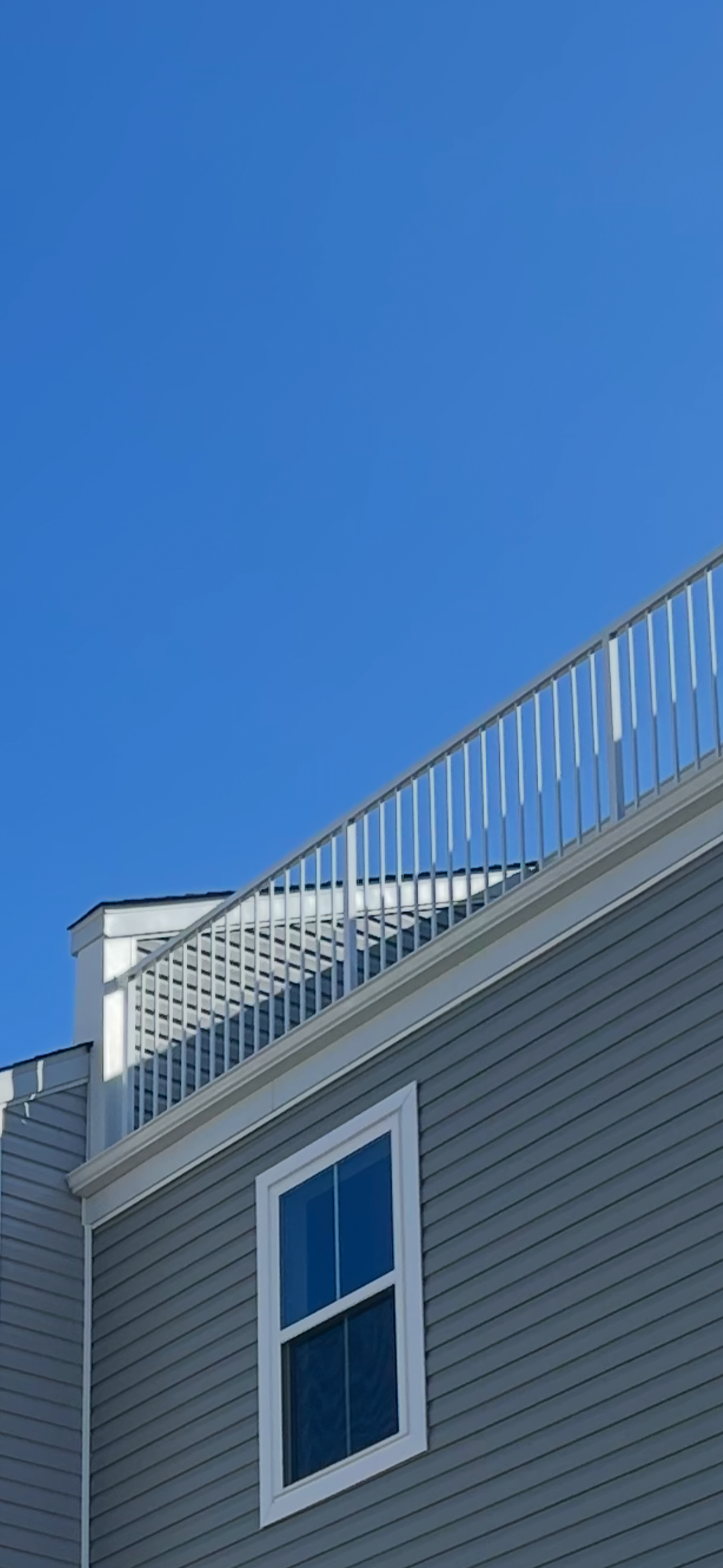 A house with gray siding, a double window, and a white railing on the balcony under a clear blue sky.