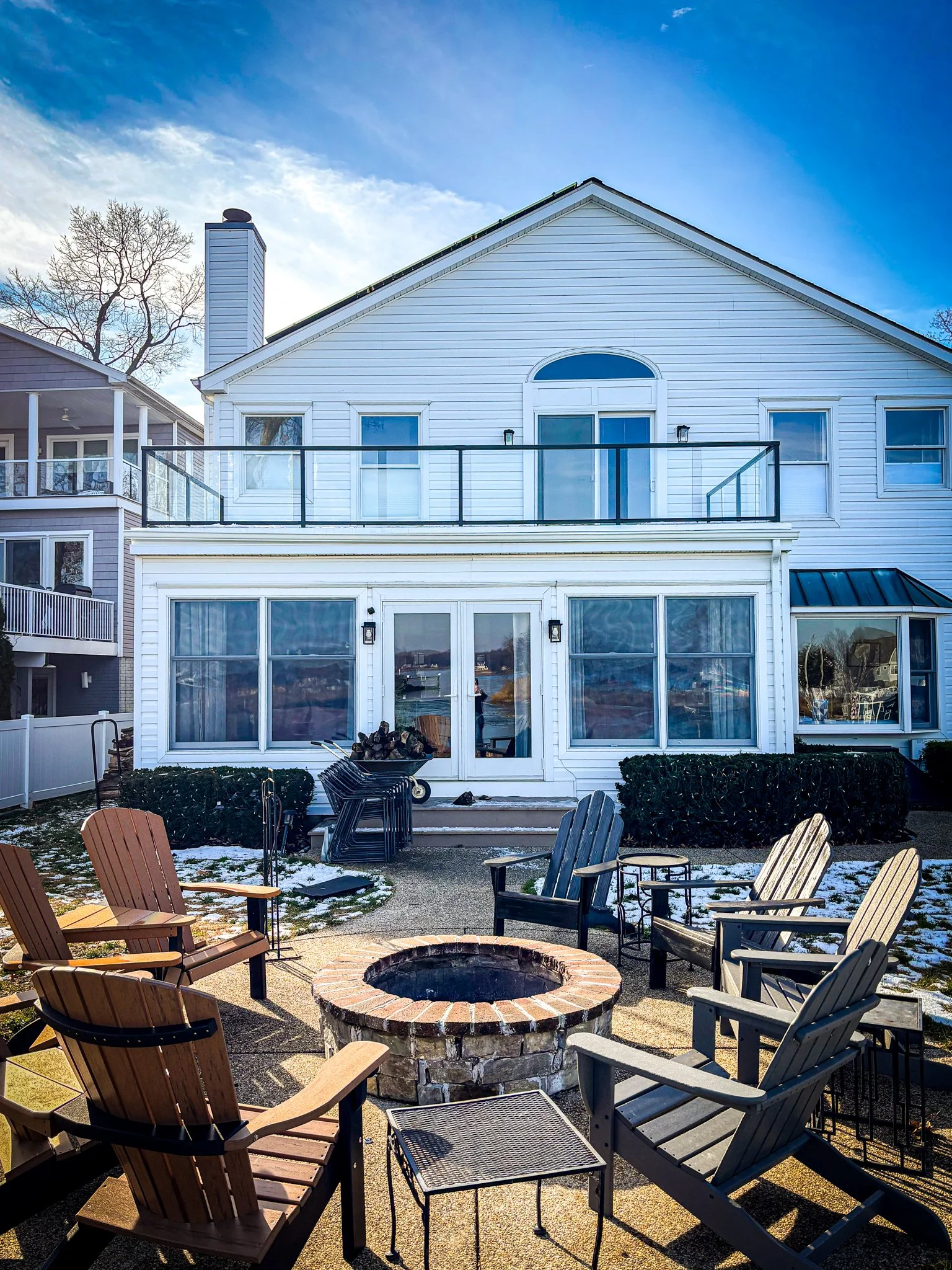 Backyard patio with Adirondack chairs arranged around a fire pit, indicating a cozy outdoor gathering space. The yard has some patches of snow, suggesting winter or early spring. The background features a two-story house with a large glass door and a