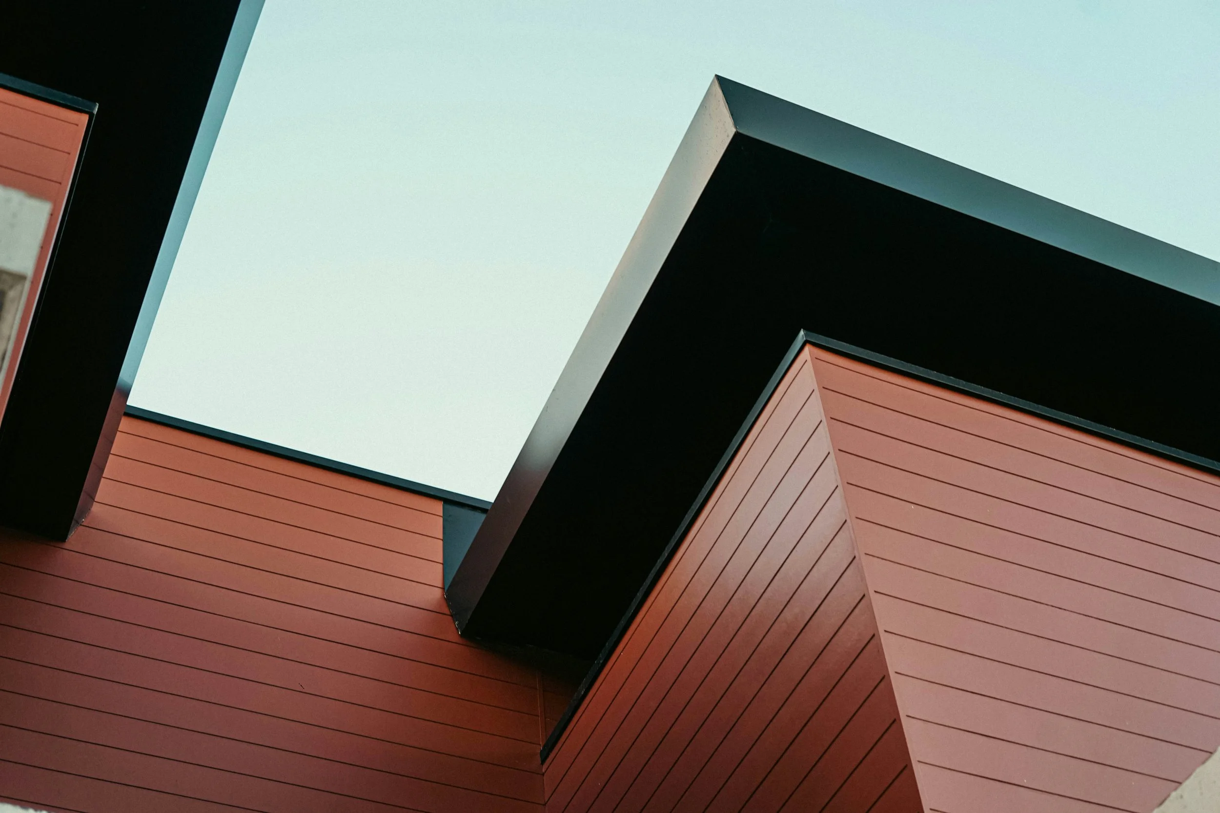 Close-up of a modern building with sharp angles, dark and reddish exterior walls, and a pale blue sky in the background.
