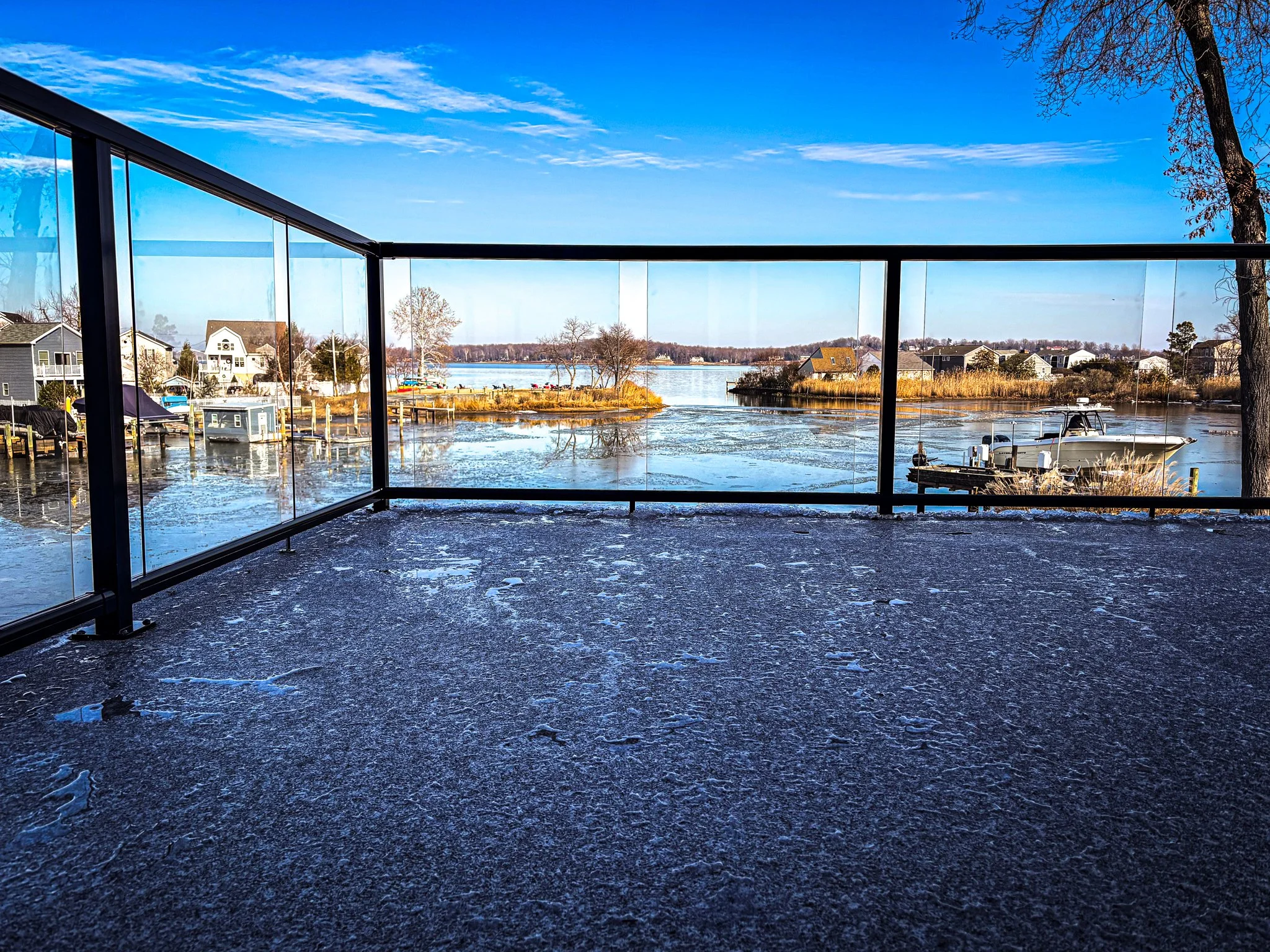 View of a lakeside neighborhood with houses, boats, and docked boats, seen through glass balcony railing on a winter day with clear blue sky.