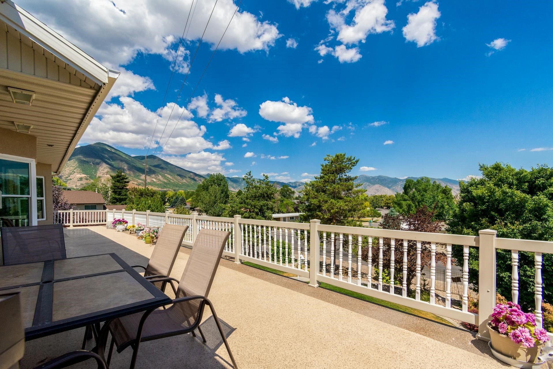 A spacious outdoor balcony or patio area with a table and chairs, surrounded by a white railing, overlooking a scenic view of mountains, trees, and a partly cloudy sky.
