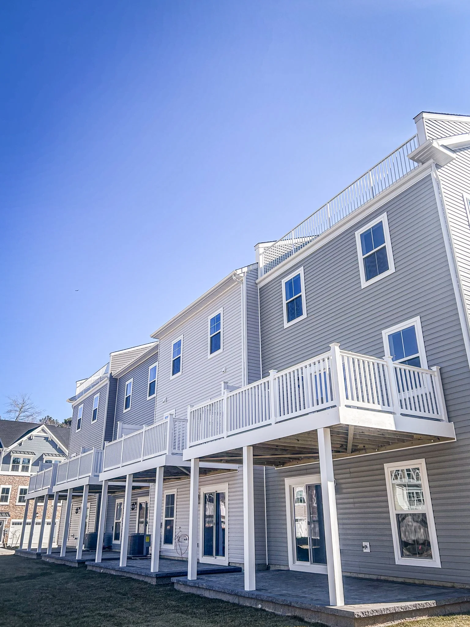 Three-story beige house with white trim and multiple large windows, featuring a wooden deck with white railings on the second floor and a covered patio underneath, under a clear blue sky.