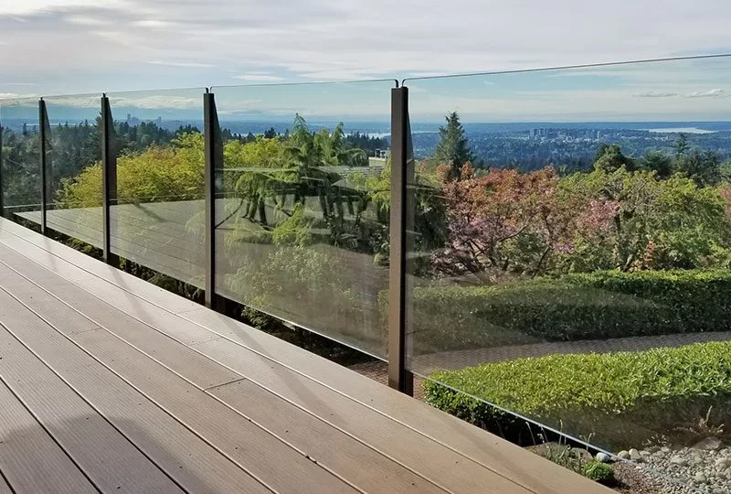 Modern outdoor deck with glass railing overlooking a lush green landscape and distant city skyline under a partly cloudy sky.