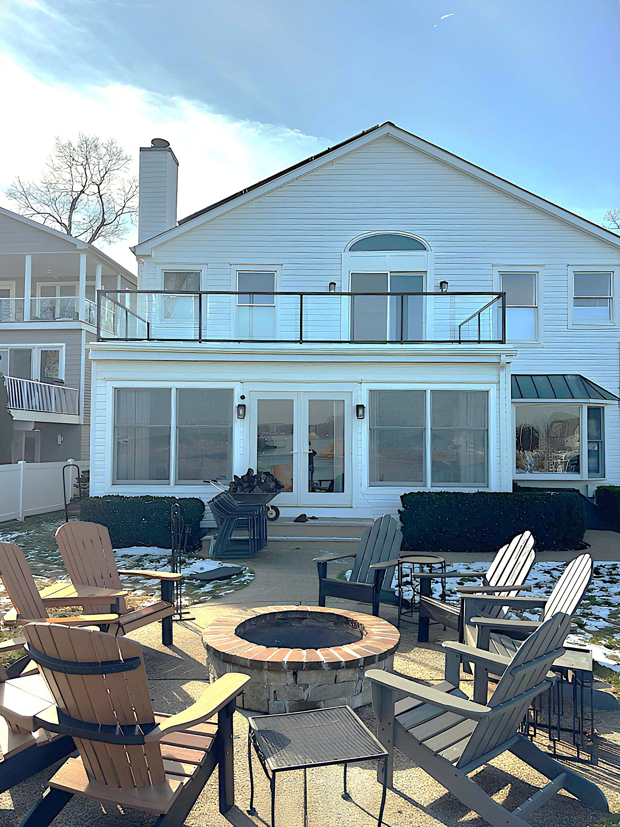 Backyard patio with Adirondack chairs arranged around a fire pit, patio is partly covered with snow, large house with multiple windows and a balcony, clear sky with some clouds.