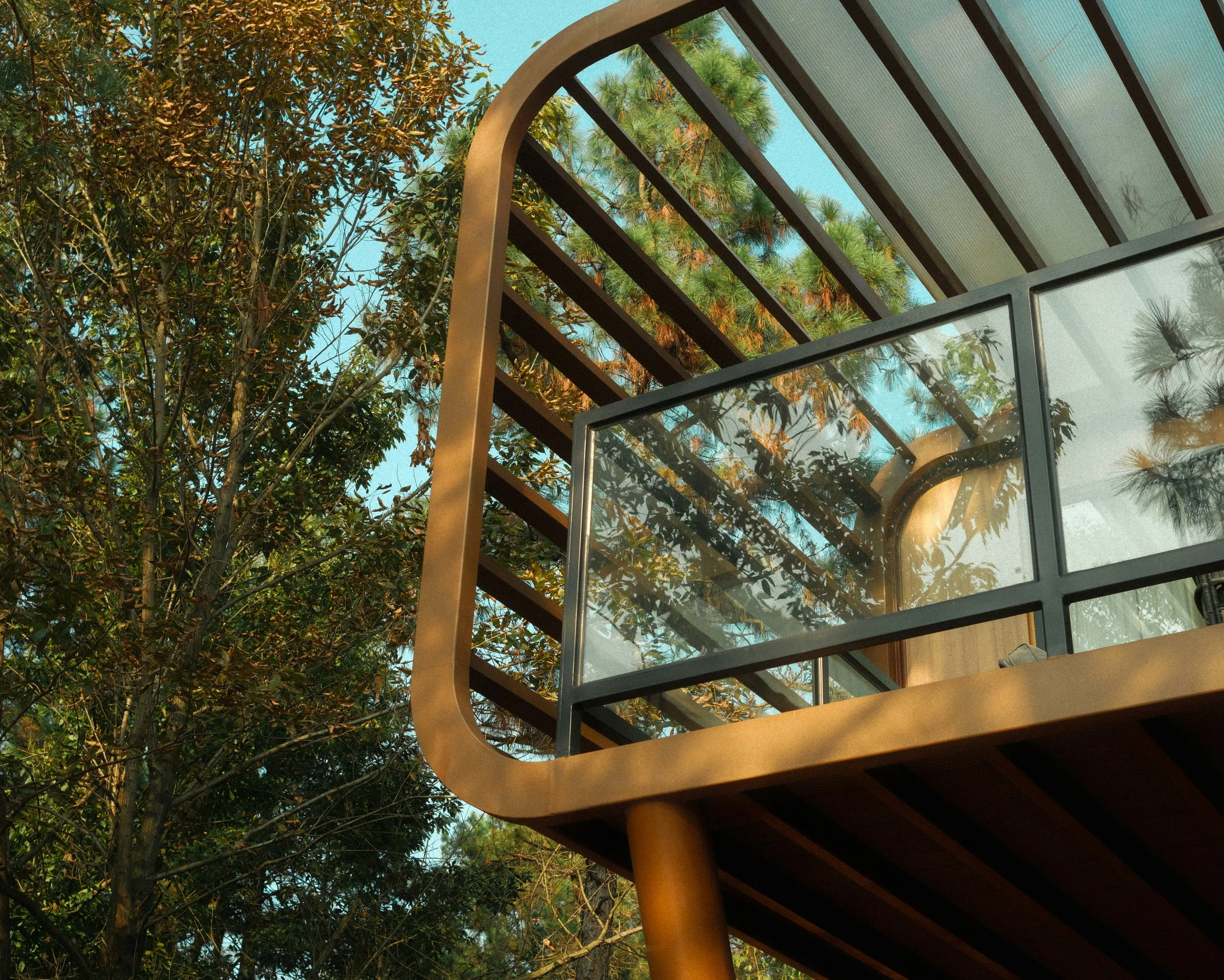 A view of a building taken from below, showing a glass balcony and a tree with green leaves in the background.
