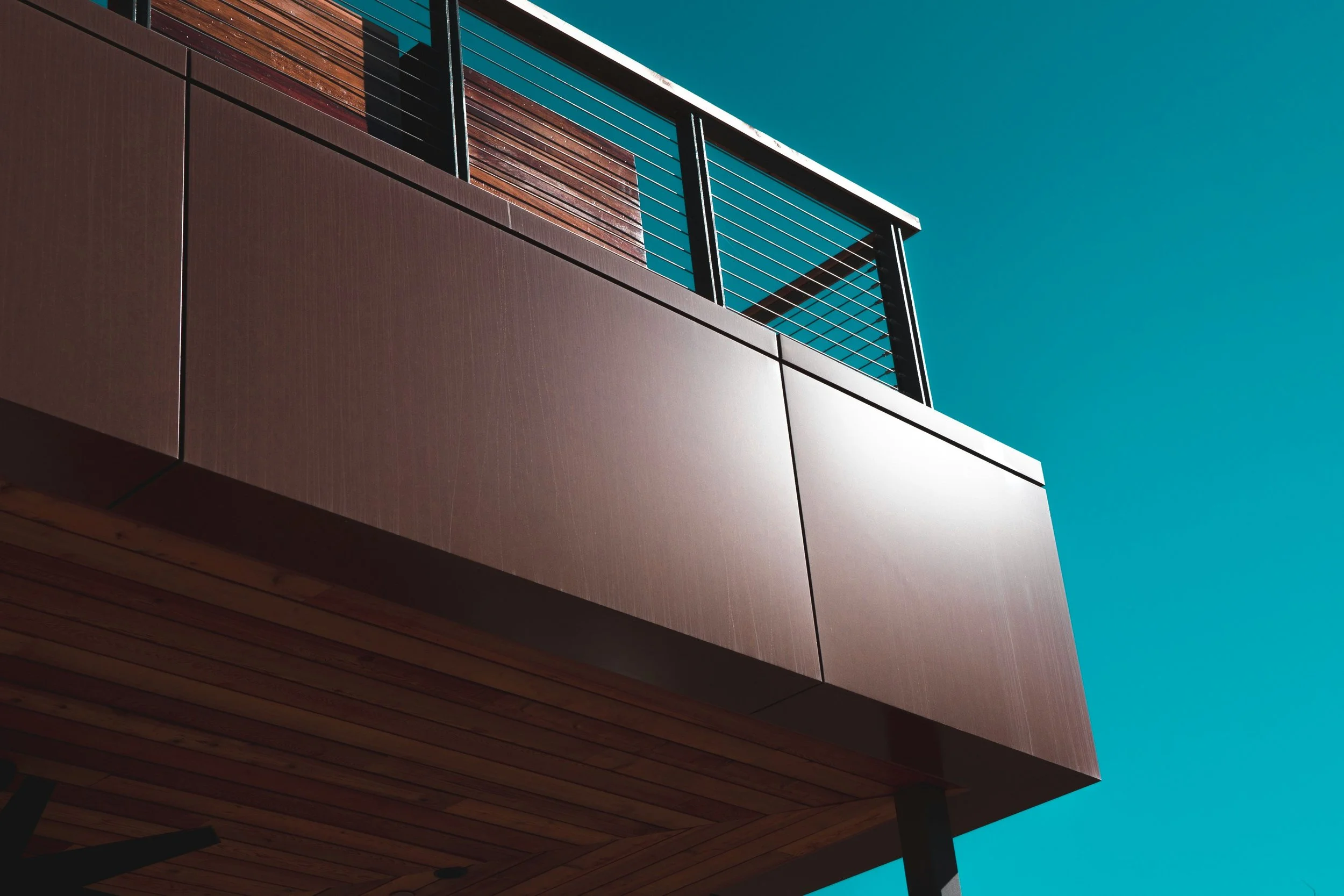 The image shows the corner of a modern building with a combination of brown, wooden, and metal elements, set against a clear blue sky. The building features horizontal and vertical lines from the construction materials and metal railings.