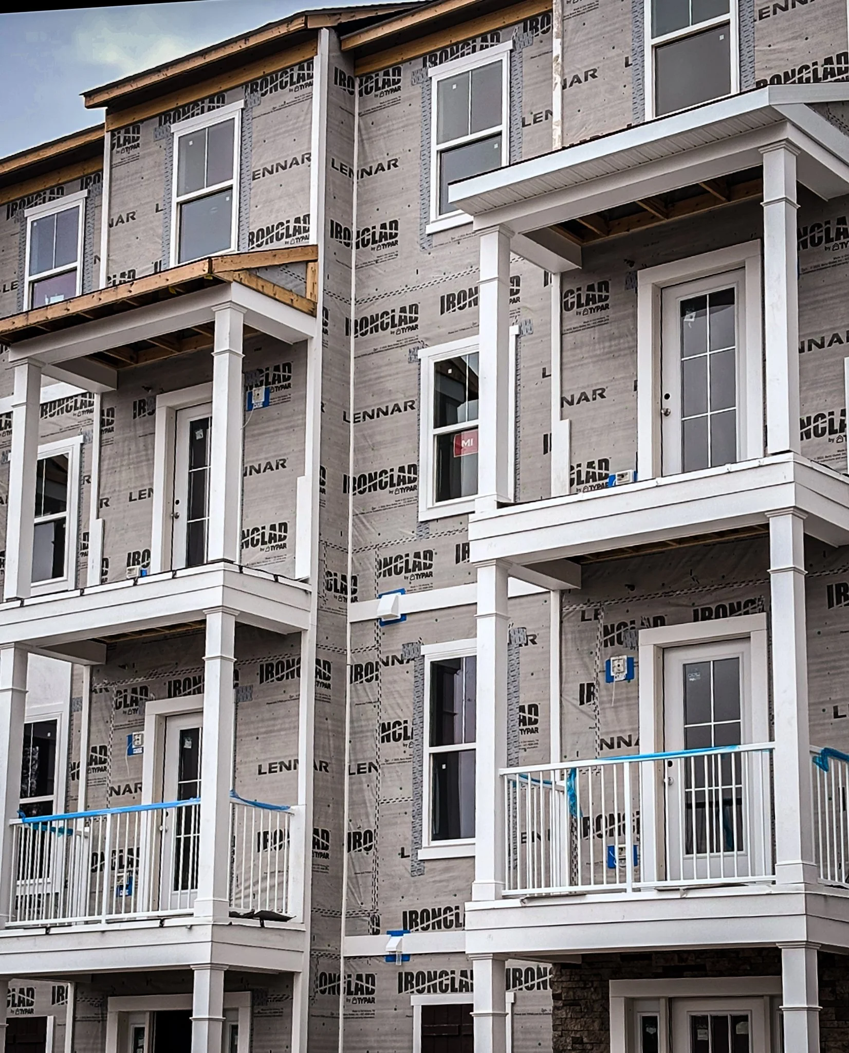 Multi-story building under construction with exposed framing, white doors, and windows. The exterior is covered with building wrap labeled 'Ironclad' and 'Lennar'.