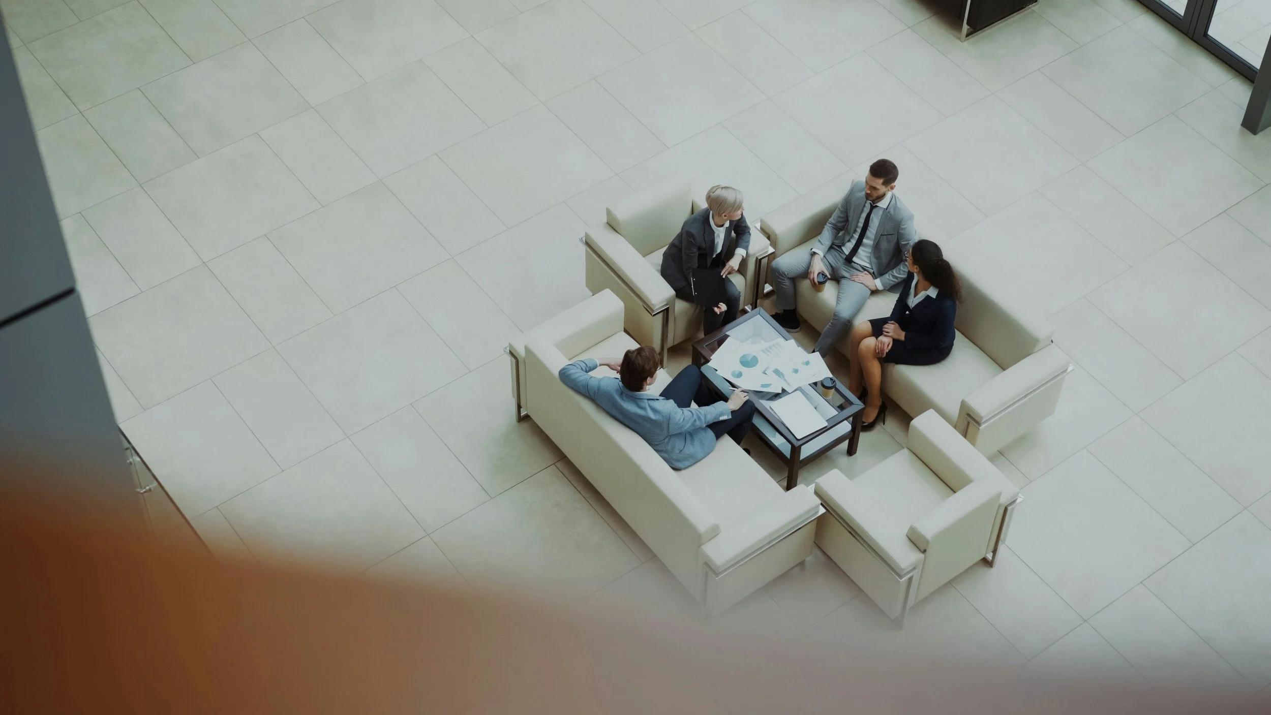 Four business professionals seated on a white sectional sofa in a modern office lobby, engaged in a discussion with papers and a tablet on a coffee table.