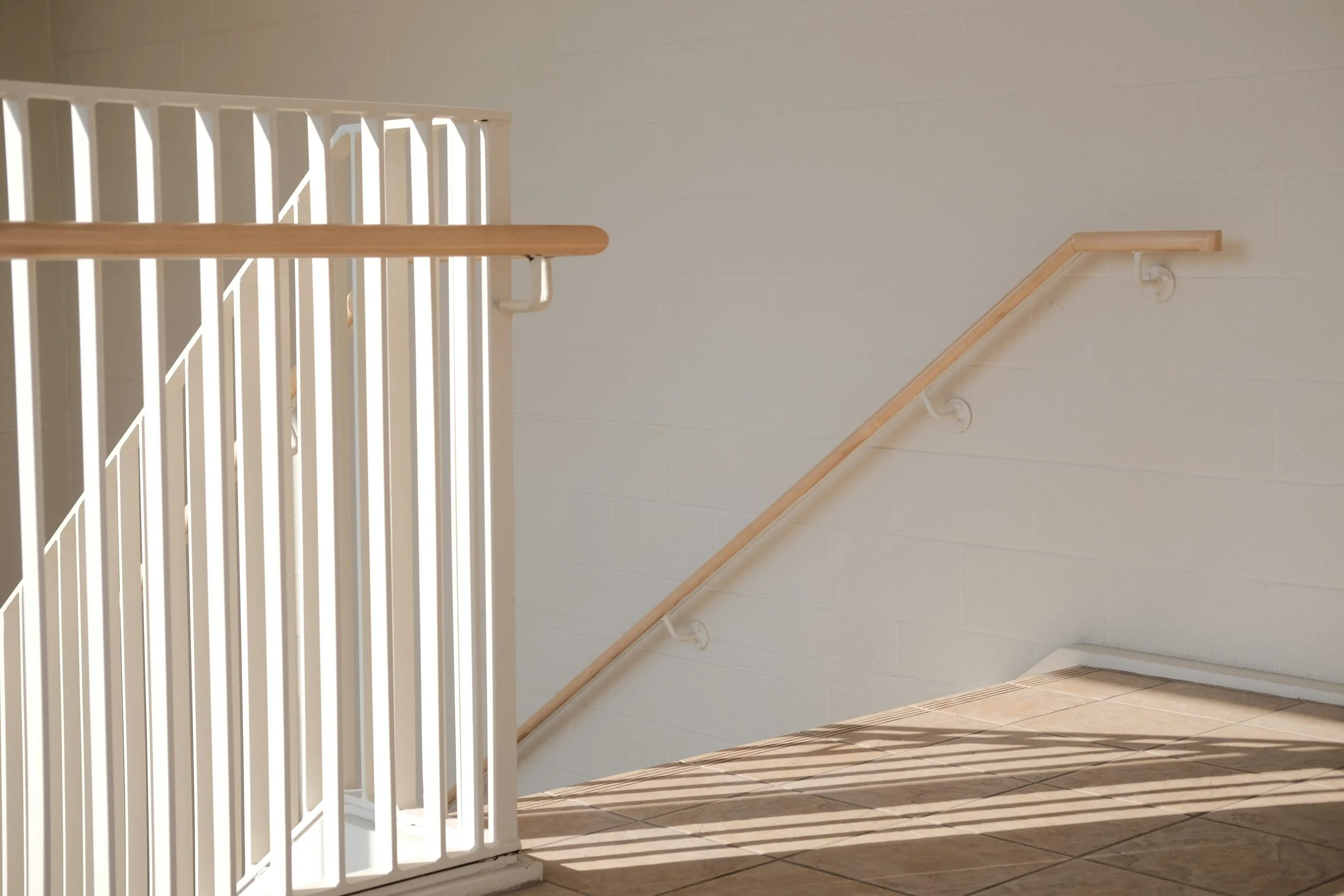 Indoor staircase with wooden handrail and white metal railing, casting shadows on the light-colored tiled floor and beige wall.
