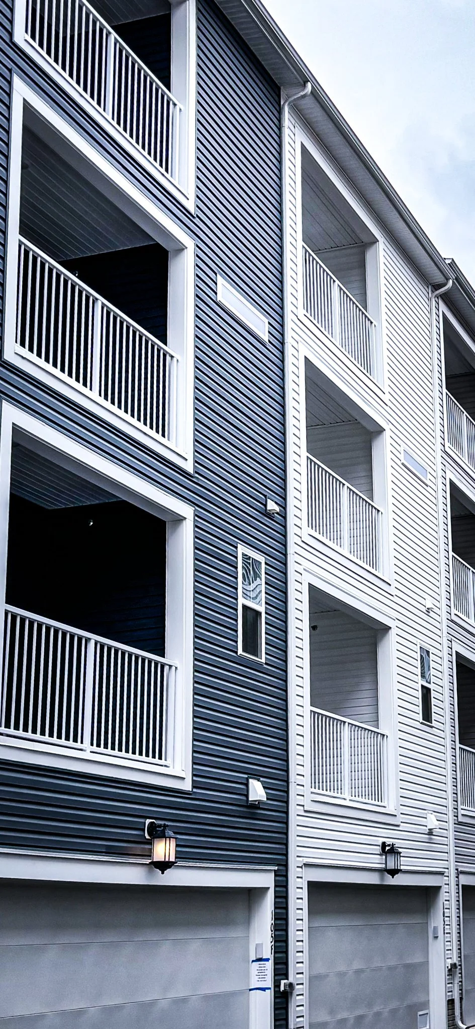 Multi-story residential building with blue and white siding, featuring balconies with white railings and light fixtures.