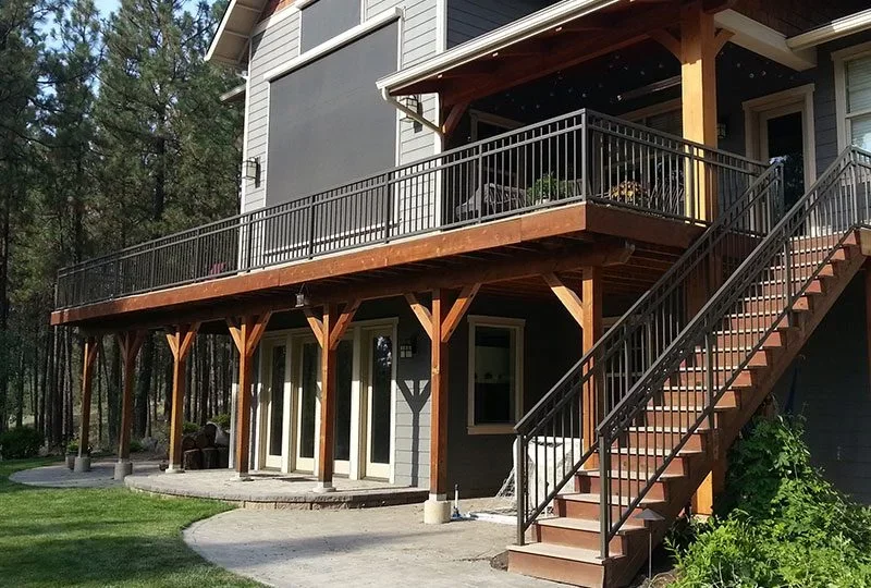 A two-story house with a wooden deck on the upper level, supported by wooden beams and enclosed with black metal railings, with stairs leading up to the deck on the right side.