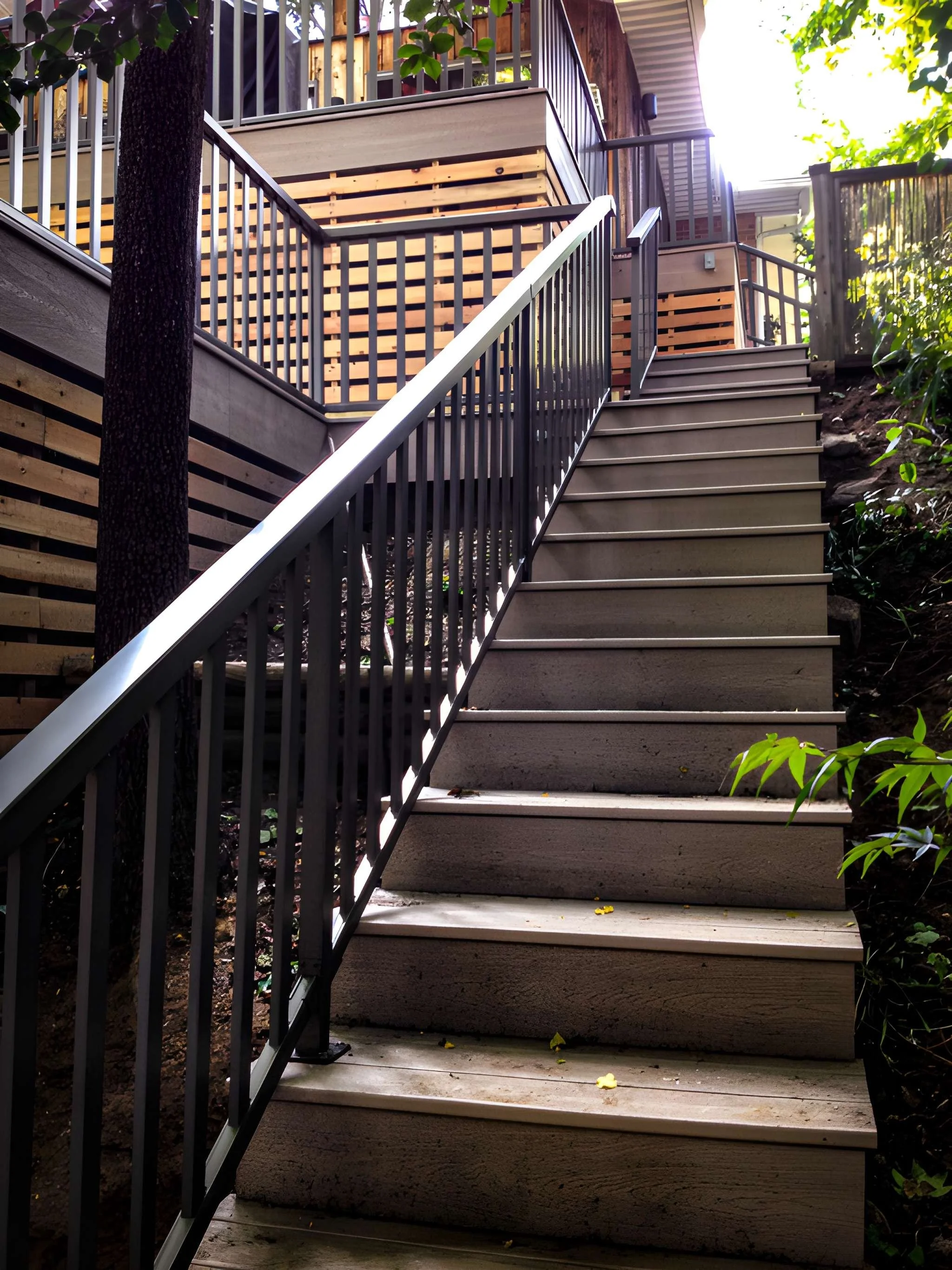 Wooden stairs leading up to a deck, with a black metal railing on both sides, surrounded by trees and greenery, sunlight shining through.