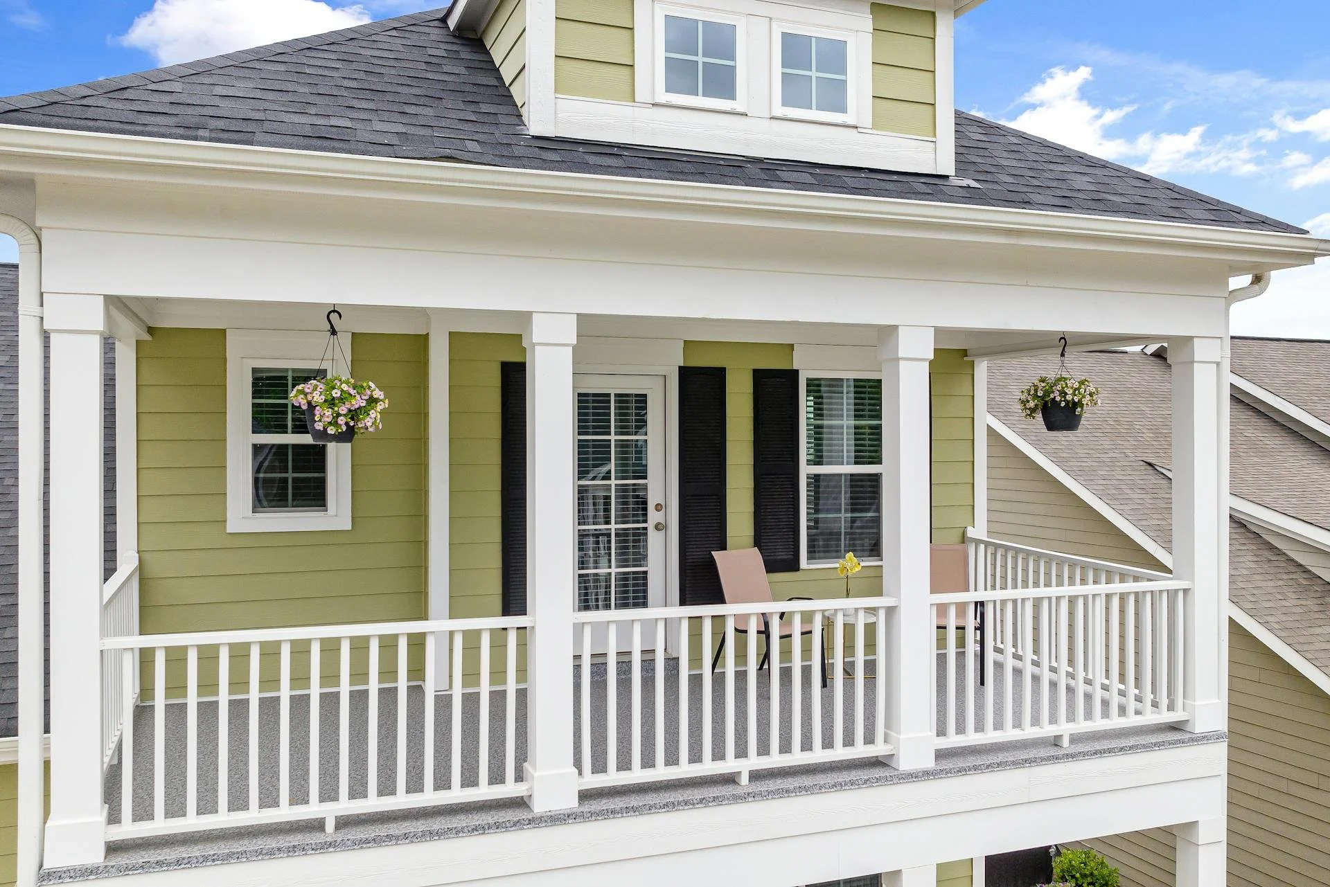 Second-story porch with white railing, hanging flower baskets, and two chairs outside a yellow house with white trim and a door with glass panels, under a gray shingled roof, against a blue sky with clouds.