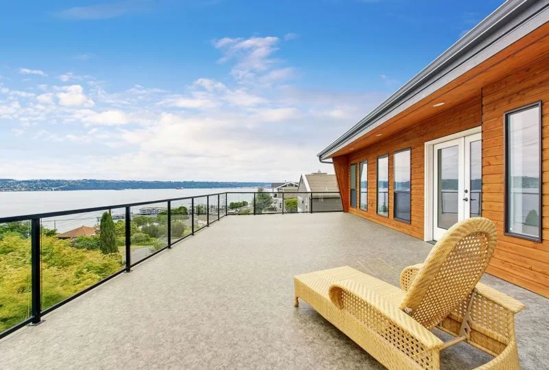 Large balcony with beige lounge chair, metal railing, ocean view, and modern wooden house exterior with large windows and doors.