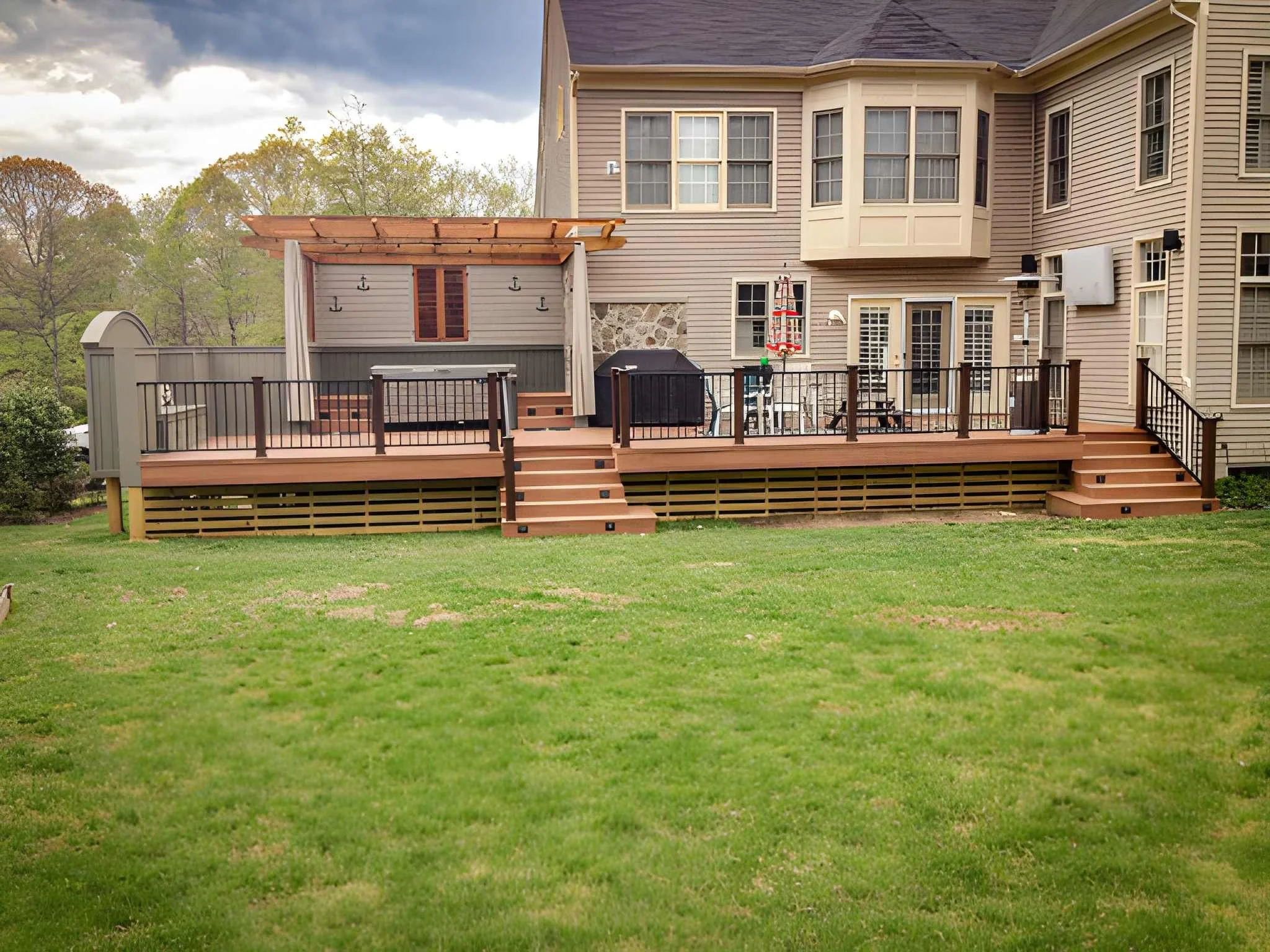 A backyard deck with black railing, stairs, and a small covered area with hooks on the wall, attached to a beige house with multiple windows, set against green trees and a cloudy sky.