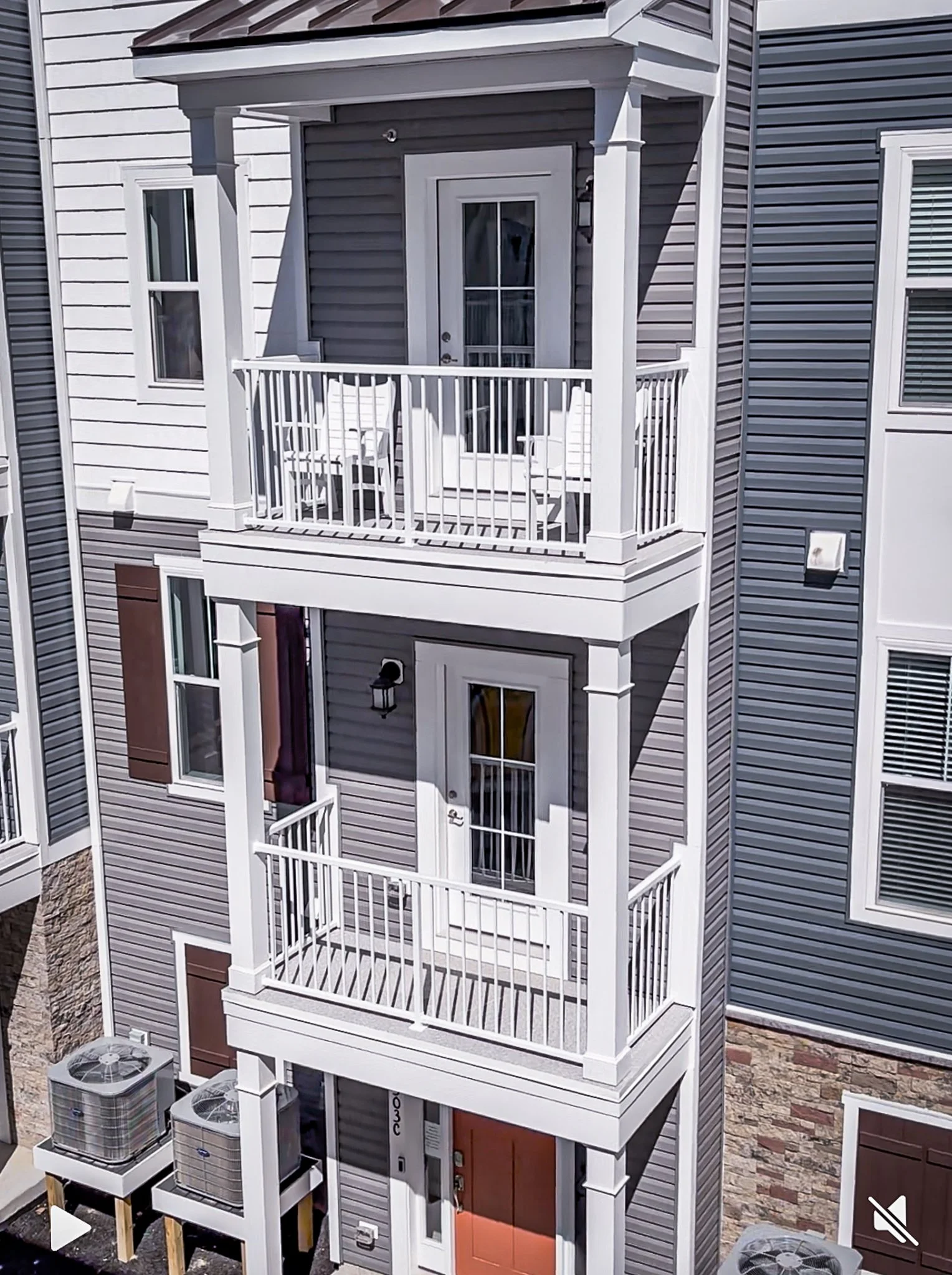 Multi-story residential building with beige and dark blue siding, featuring multiple balconies with white railings, and a ground-level entrance with a reddish-brown door. Two air conditioning units are visible at the bottom.