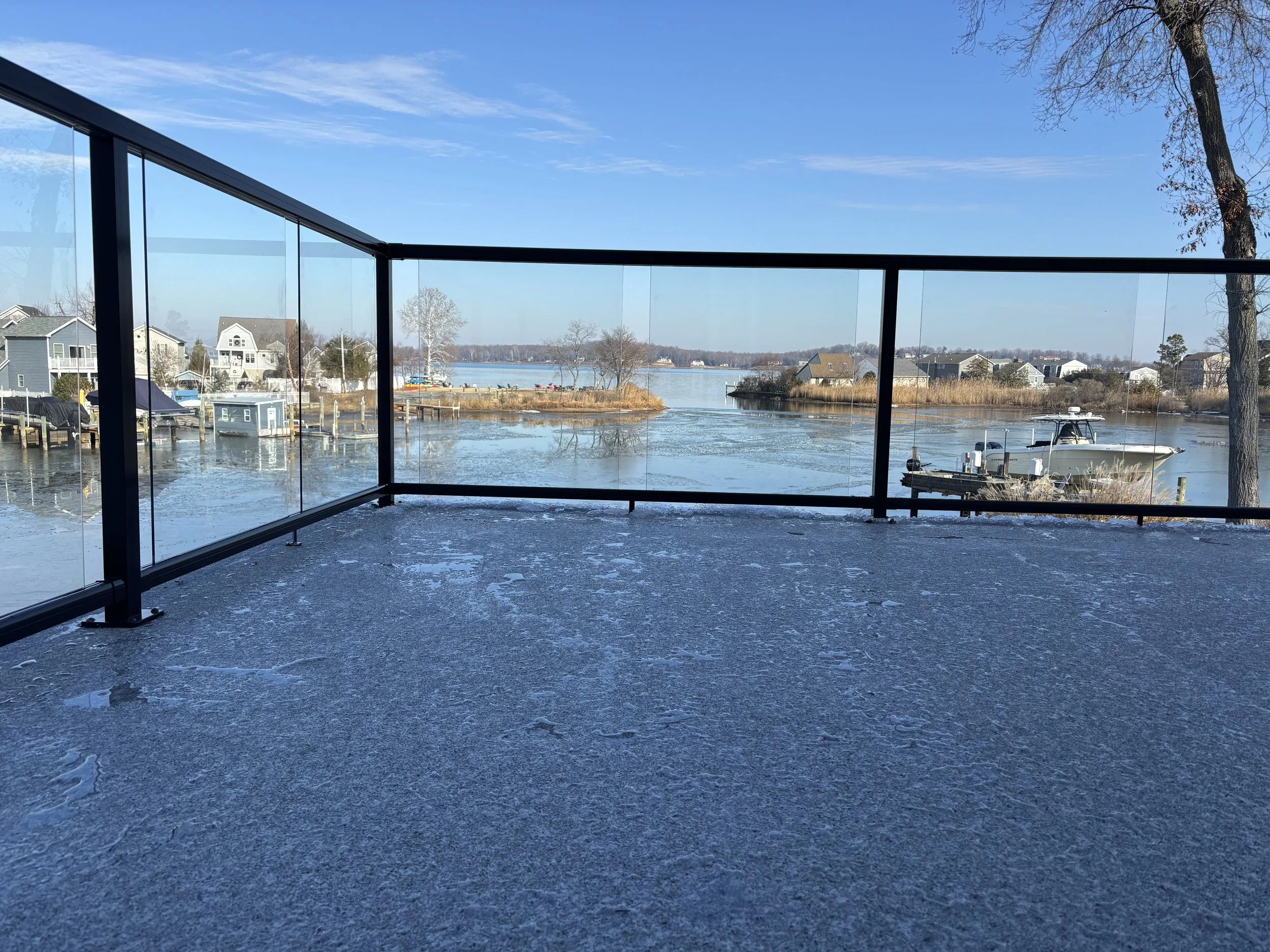 View of a house and boats on a waterfront as seen from a balcony with glass panels, with a tree on the right and a partly icy deck in the foreground.