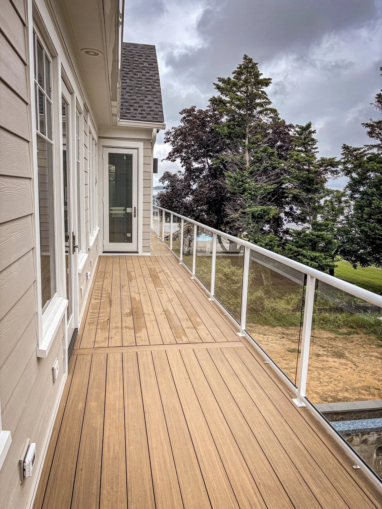 A long balcony with wooden decking, white railing with glass panels, beige house exterior with multiple windows and a door, tall trees and cloudy sky in the background.