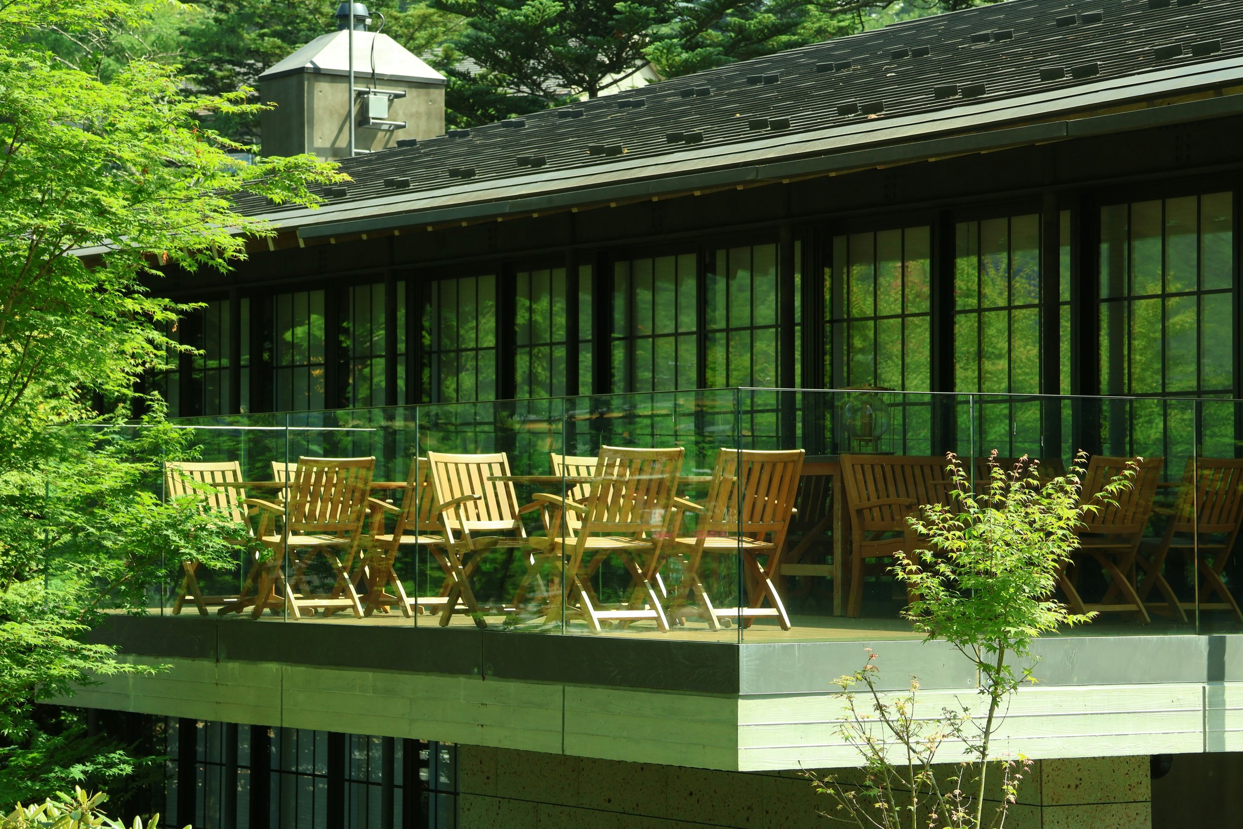 A modern building with large glass windows, a black tiled roof, and surrounding lush green trees and bushes.
