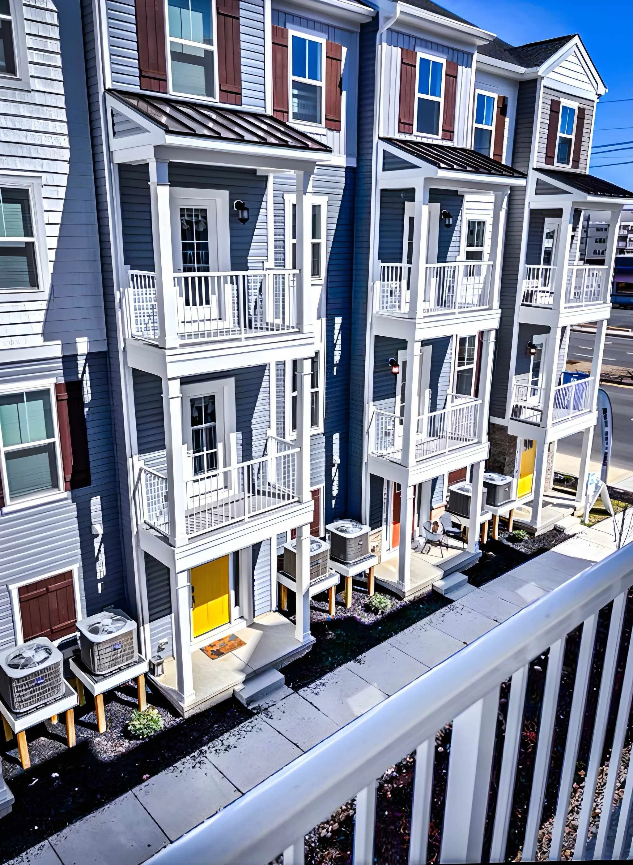 Modern townhouse with multiple balconies, white railings, and blue siding, with some units featuring brown window shutters and yellow door accents, seen from a balcony with a white railing in the foreground.