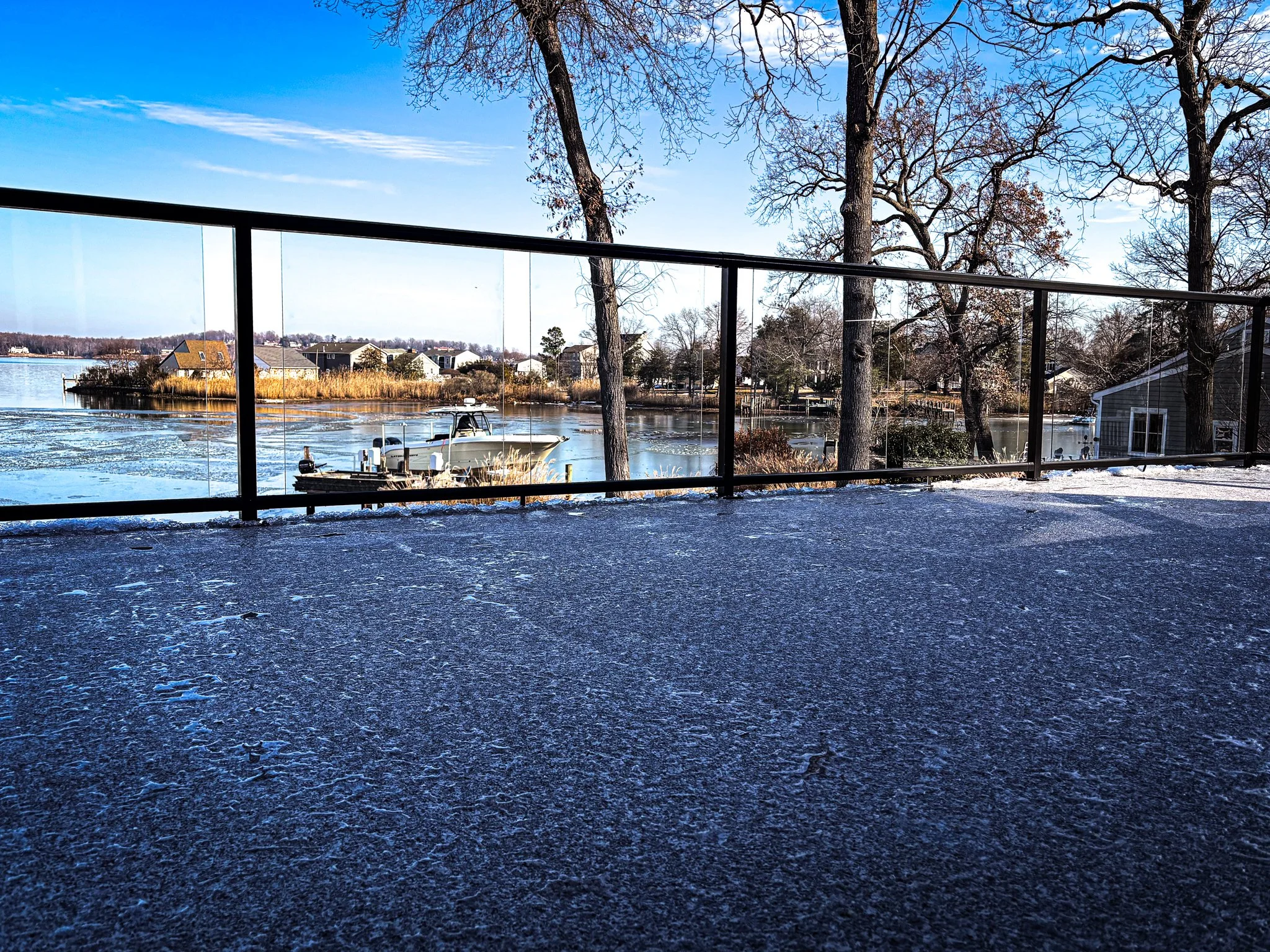 Scenic view through a glass railing of a lake with boats, trees with bare branches, some houses on the shoreline, and a partly frozen surface under a clear blue sky.