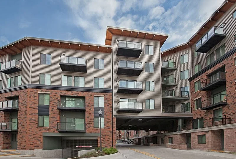 Multi-story residential apartment building with brick and siding exterior, featuring balconies and a covered parking area underneath, under a partly cloudy sky.