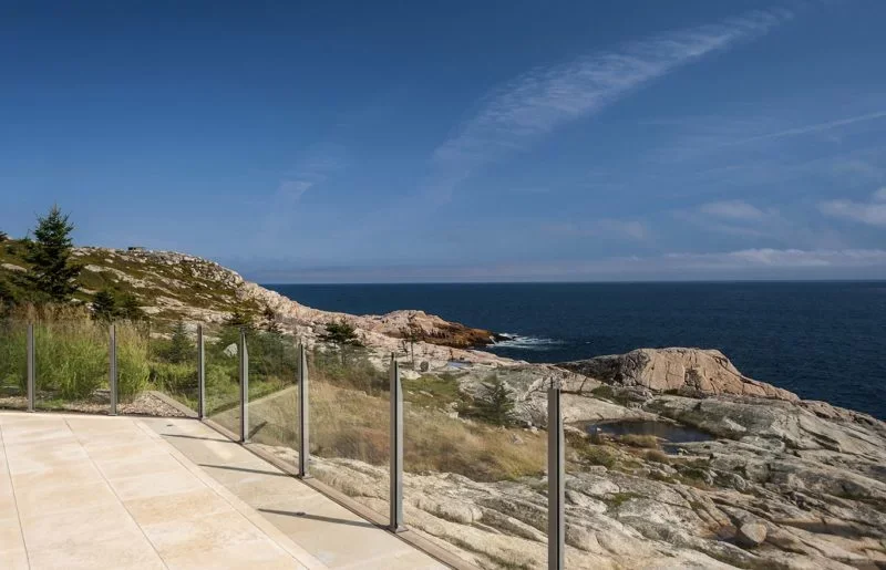Seaside view with rocky coast, grassy terrain, and ocean in the distance, seen from a balcony with glass railings on a sunny day.