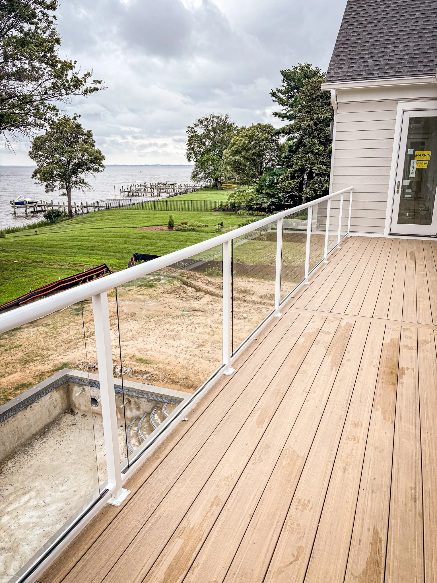 View from a balcony overlooking a grassy yard, trees, and a body of water with a dock and boat, under cloudy skies.
