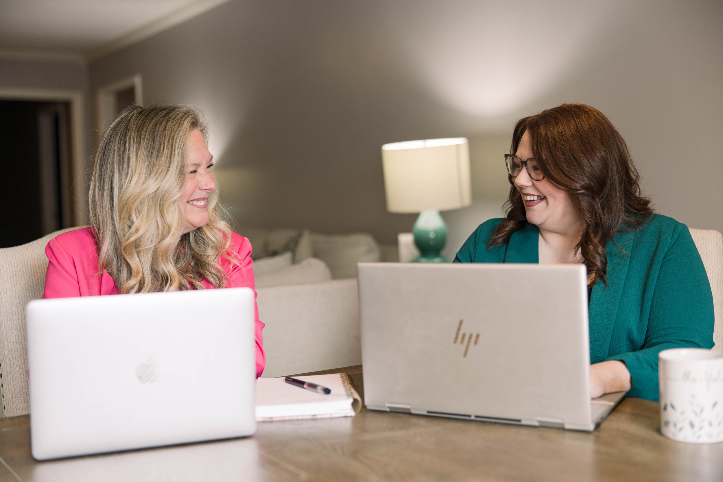 Two women sitting at a table with laptops, smiling and looking at each other, in a warmly lit room.