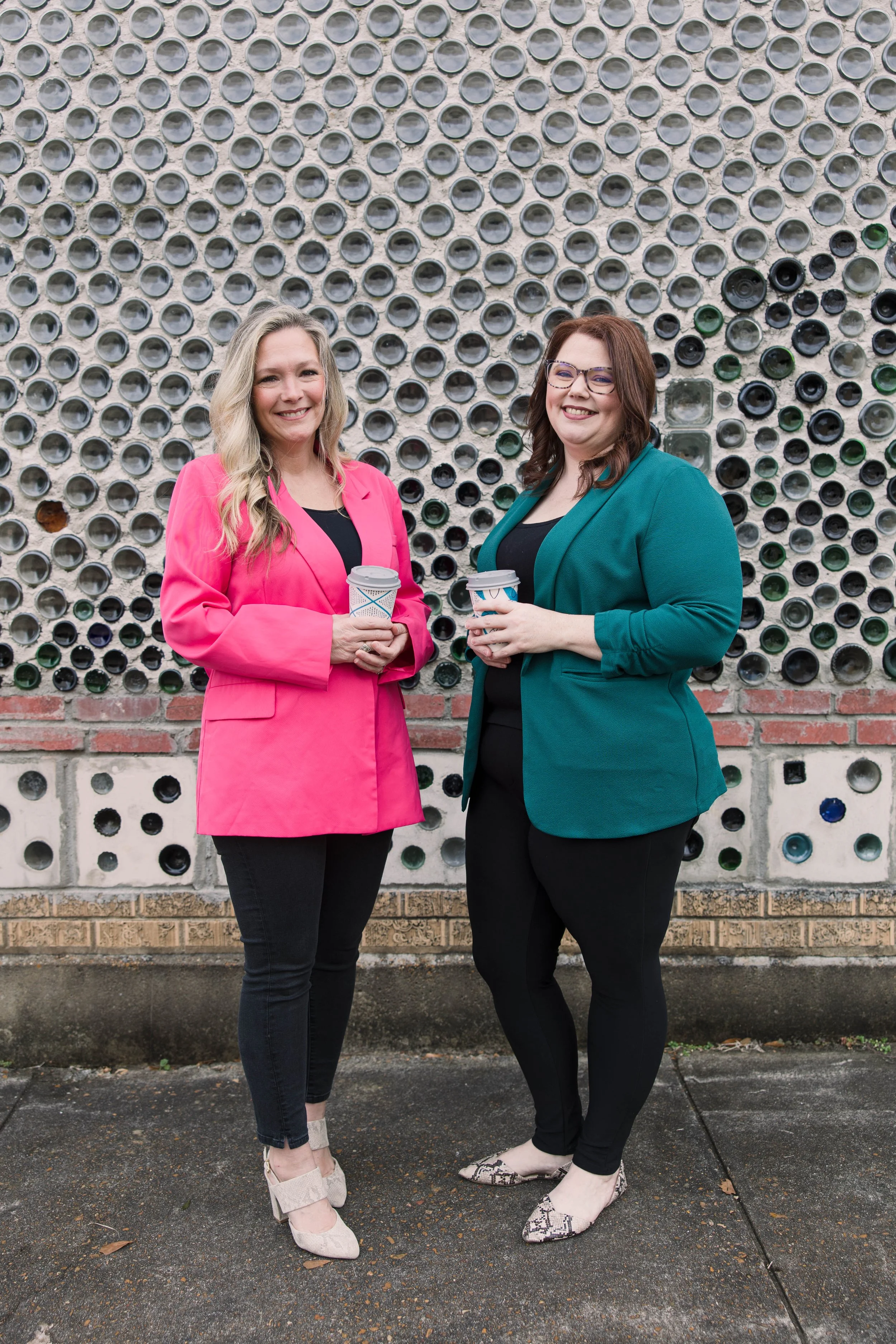 Two women standing outdoors, smiling, holding coffee cups, in front of a wall decorated with glass bottles and bricks.