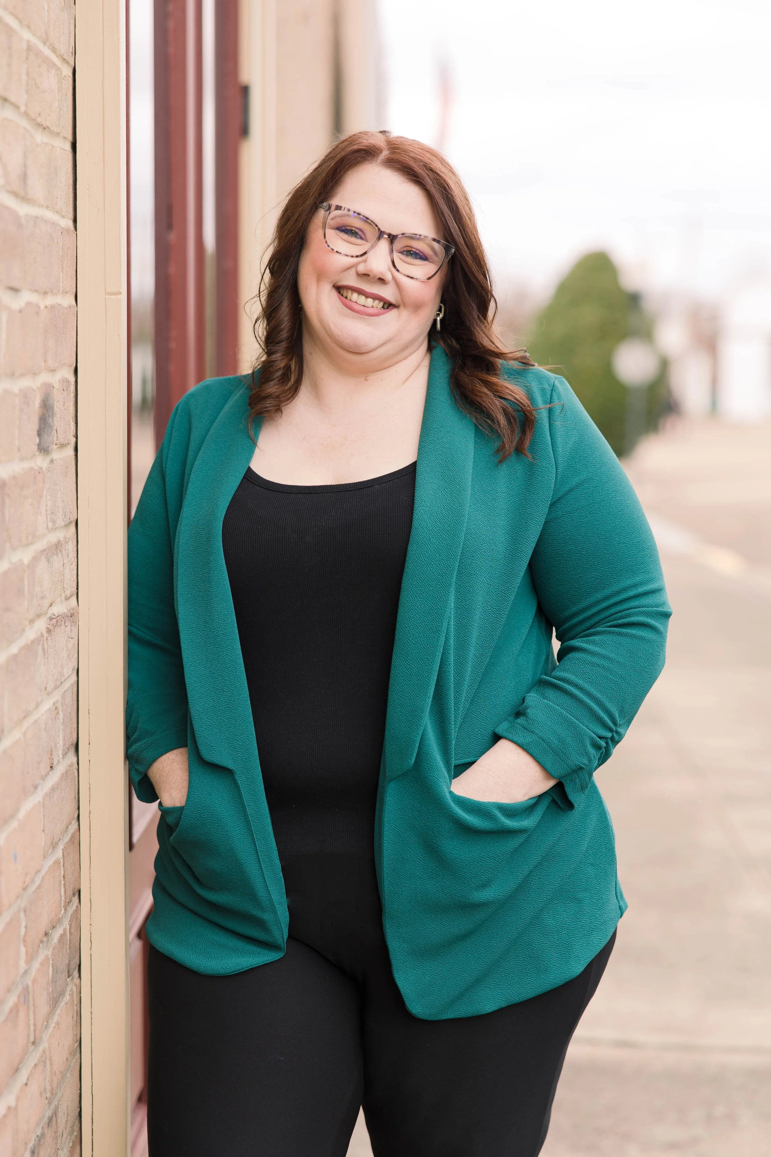 A smiling woman with red hair and glasses, wearing a teal blazer over a black top, standing outdoors on a sidewalk.