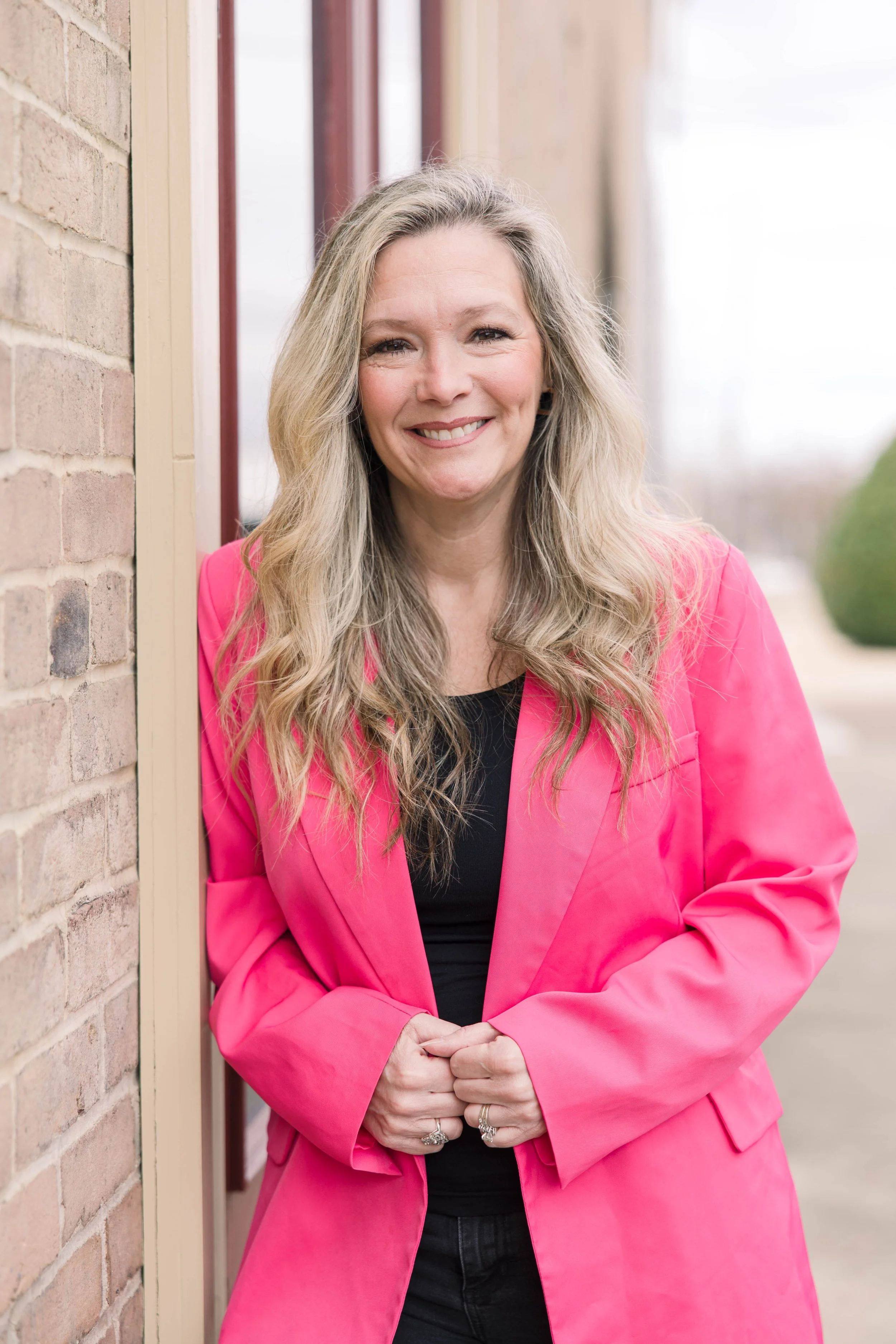 A woman with long, wavy blonde hair smiling, wearing a bright pink blazer and black top, standing outside near a brick wall and window.
