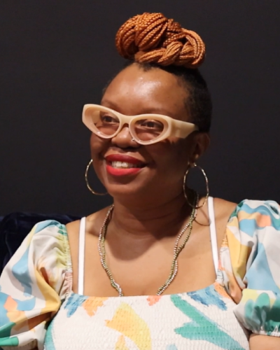 Headshot of Sabine smiling. She is wearing big bold beige glasses, bright red lipstick, and a vibrant pastel colored dress with blues, yellows, teal, and creme. Her hair is braided, colored brunette, and is tied neatly in a bun on the top of her head