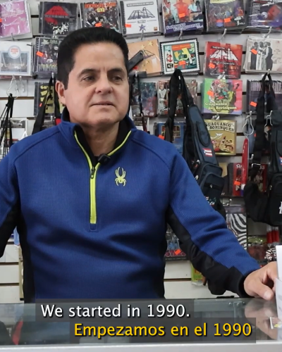 Reinaldo standing behind the counter inside El Barrio Music Center. In the background is a wall full of CDs with vibrant CD covers. He has short hair and is wearing a navy blue sweater.