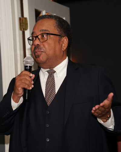 Glenn standing in front of a doorway holding a mic. He is wearing glasses and a suit with a tie. He is in the middle of saying something and his left hand is gesturing and suggestive.