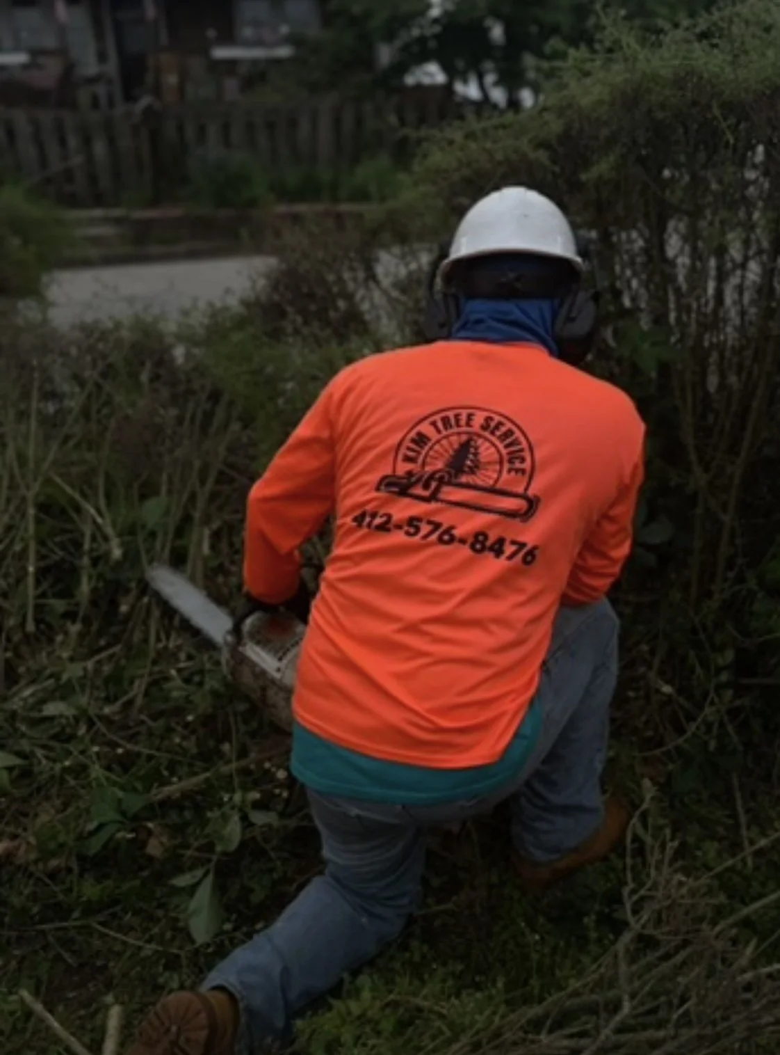 A person wearing an orange shirt with the logo 'Russum Tree Service' and a phone number, cutting a tree branch with a chainsaw in a yard with bushes.