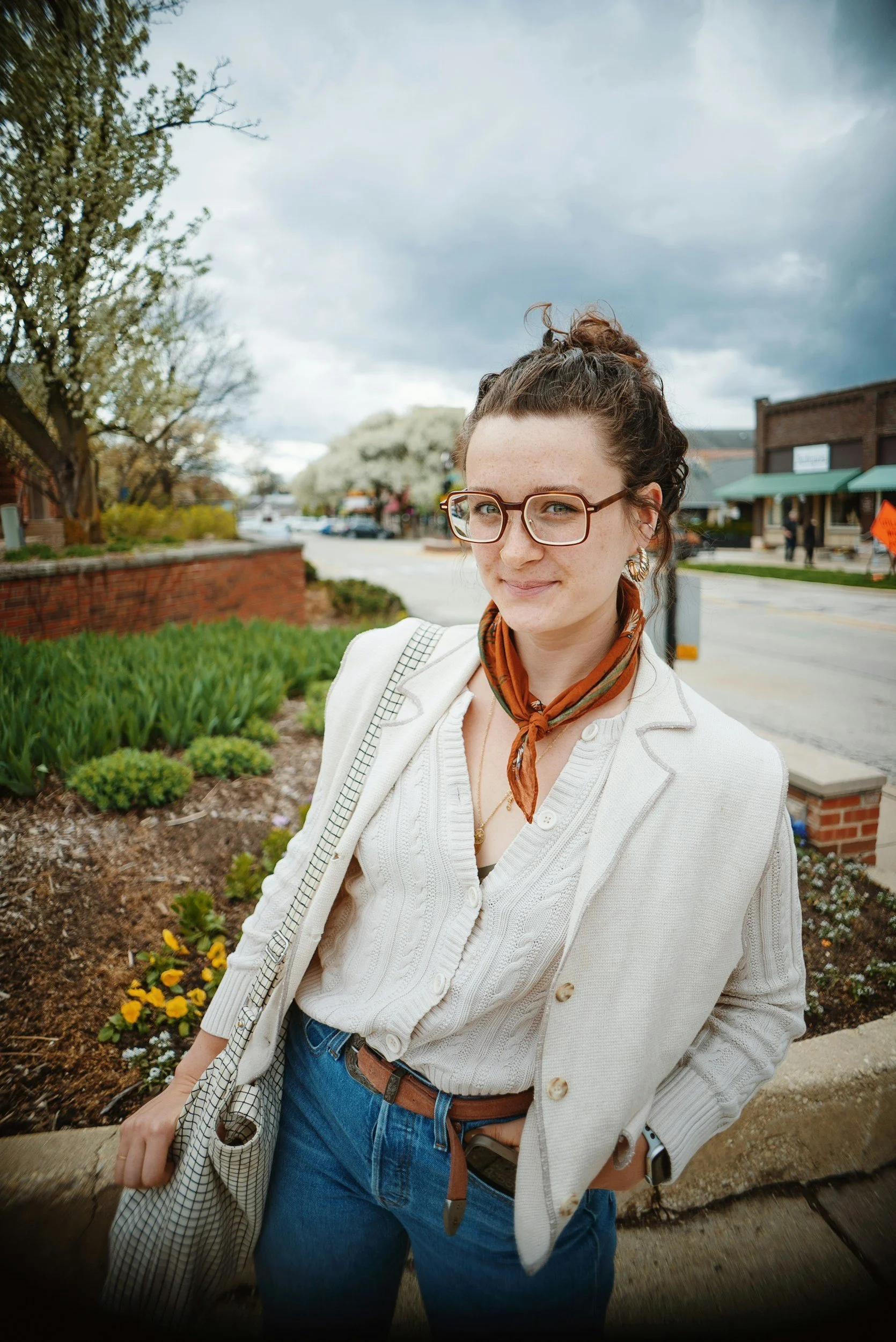 A young woman with curly hair in a bun, wearing glasses, a white sweater, and a brown bandana around her neck, standing outdoors in a small town with trees, buildings, and a cloudy sky in the background.