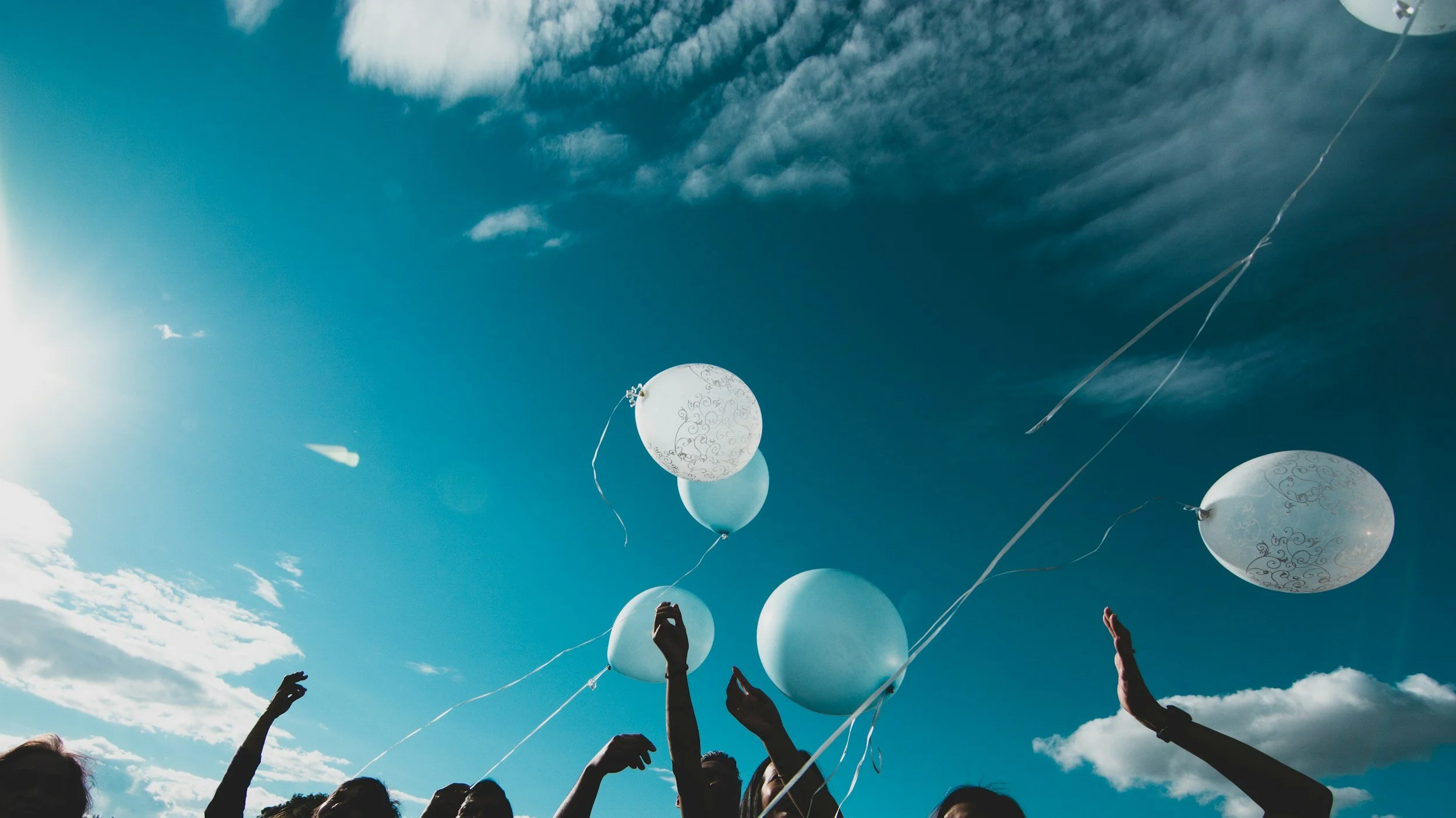 People releasing white balloons into the sky during daylight. Some hands are visible reaching upward beneath a partly cloudy sky.