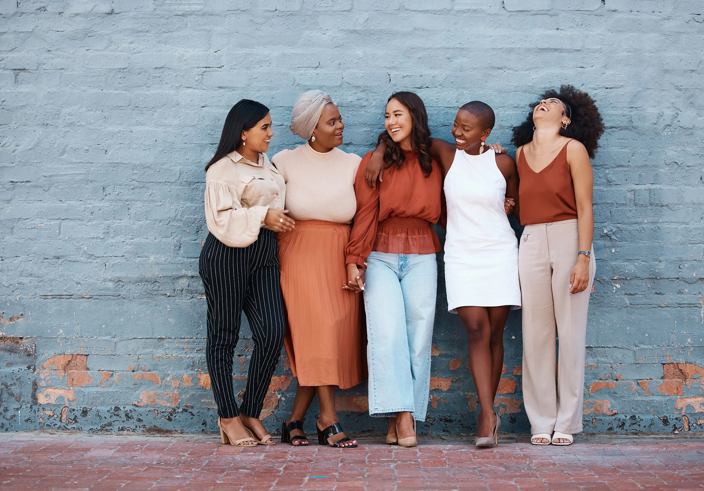 Five women standing together against a textured light blue brick wall, laughing and smiling, with one woman with her arm around another.