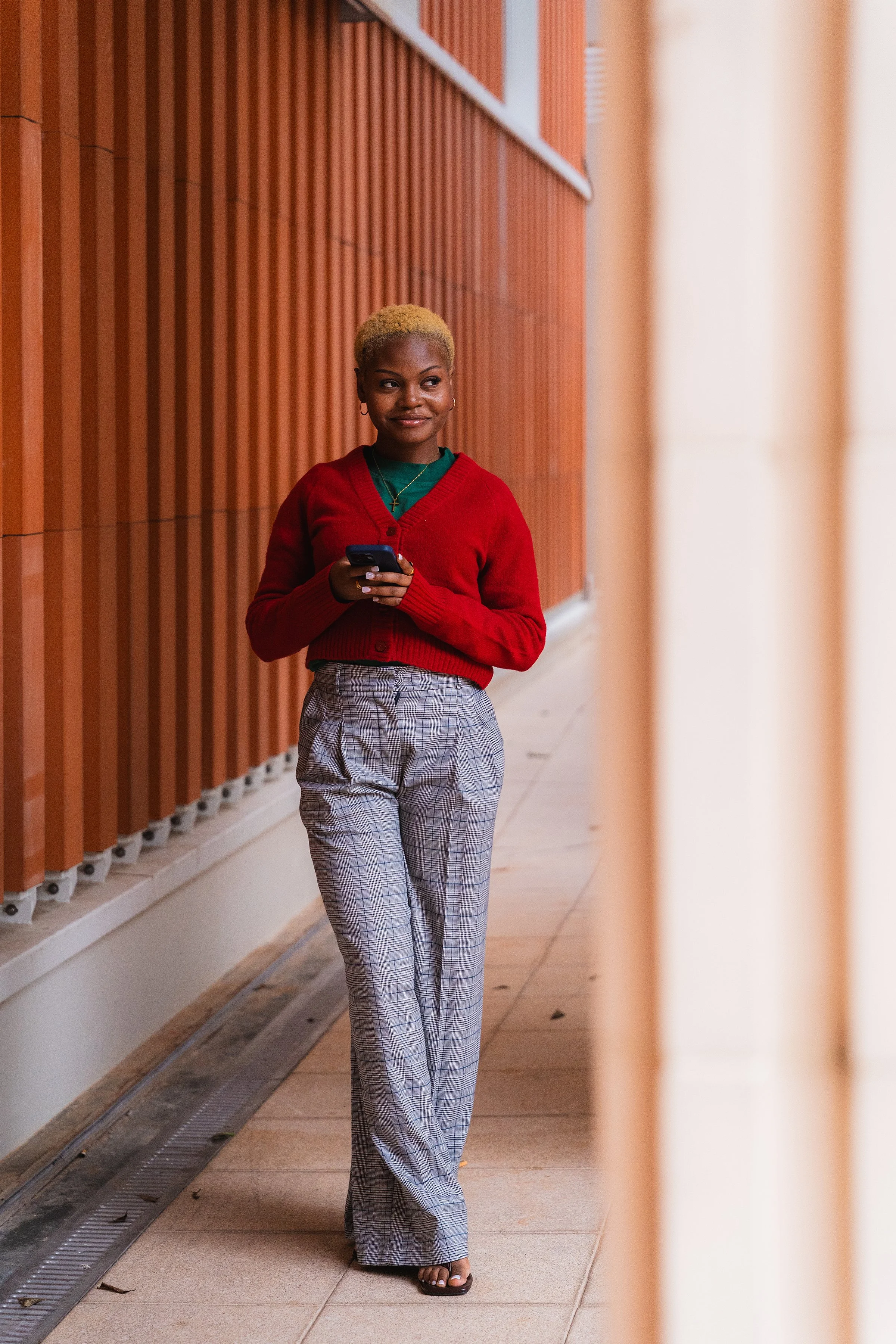A woman with short blonde hair wearing a red cardigan, green shirt, and plaid pants is standing in a hallway with wooden paneling and holding a smartphone. She has jewelry including earrings, a necklace, and a cross pendant. She is smiling slightly and looking to her left.