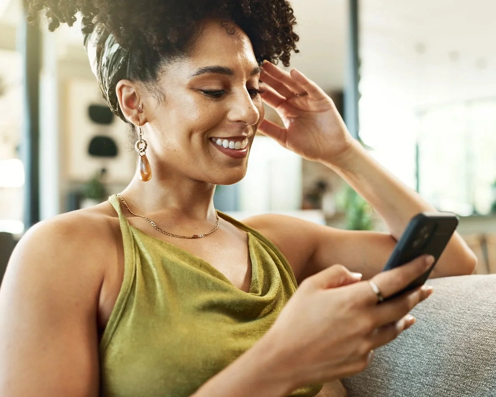 A woman with curly hair wearing a green sleeveless top and jewelry, smiling while looking at her phone in a well-lit indoor space.