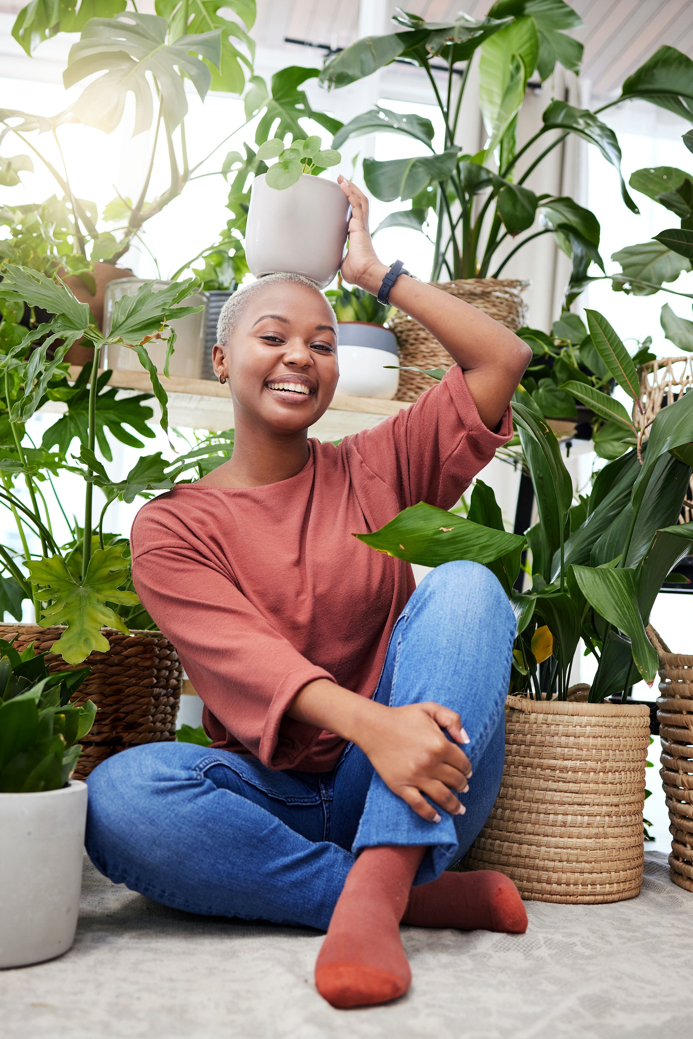 Smiling woman sitting on the floor surrounded by houseplants, holding a potted plant on her head and looking at the camera.