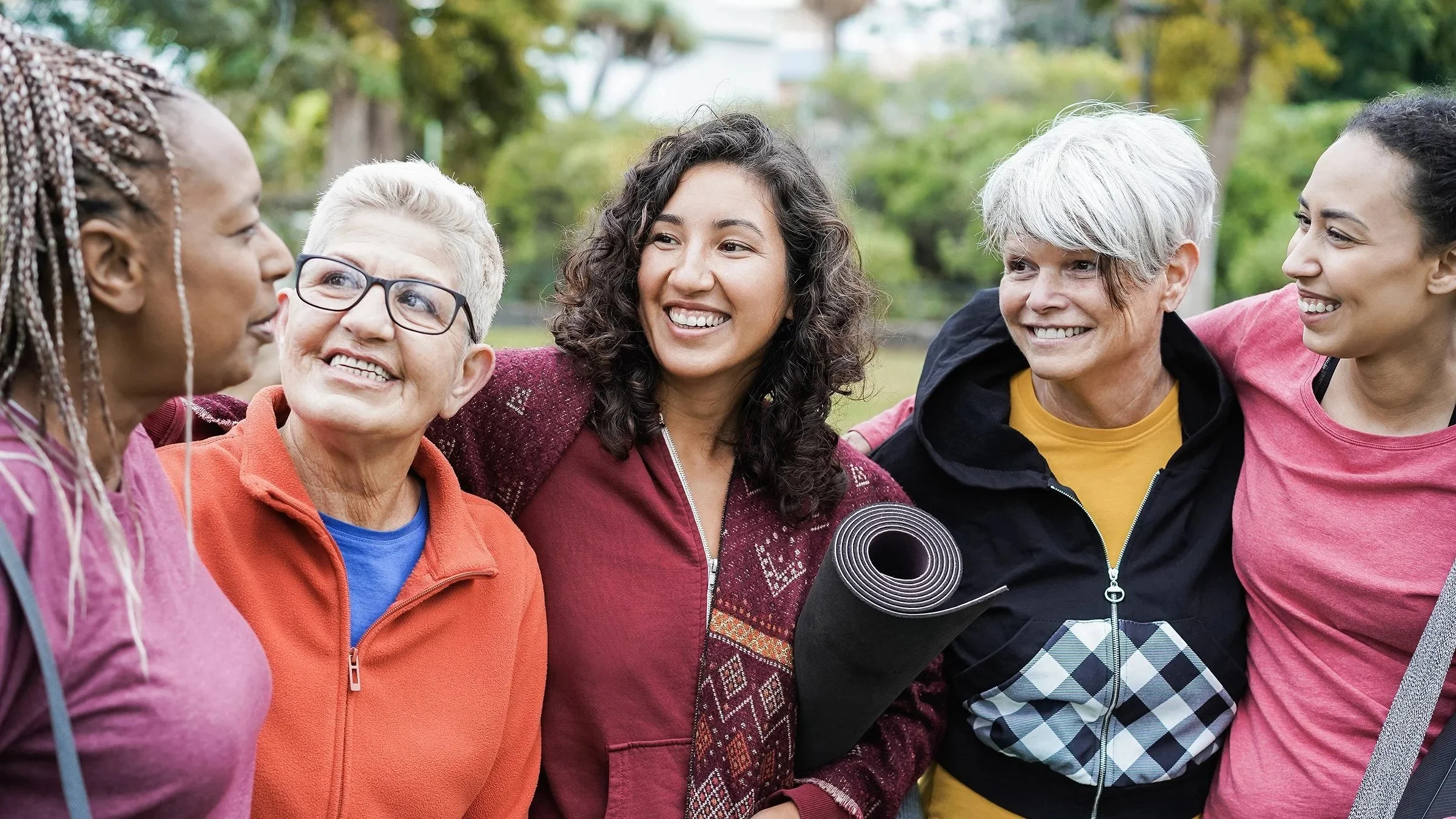 Group of five women standing together outdoors with arms around each other, smiling and talking, with trees and greenery in the background.