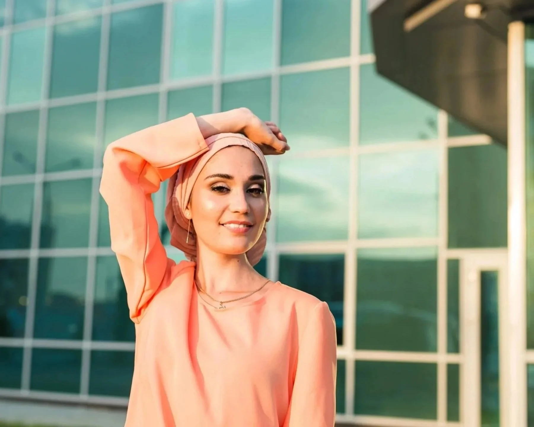A woman wearing a peach-colored shirt and a matching headscarf outdoors in front of a glass building, smiling with one hand on her head.