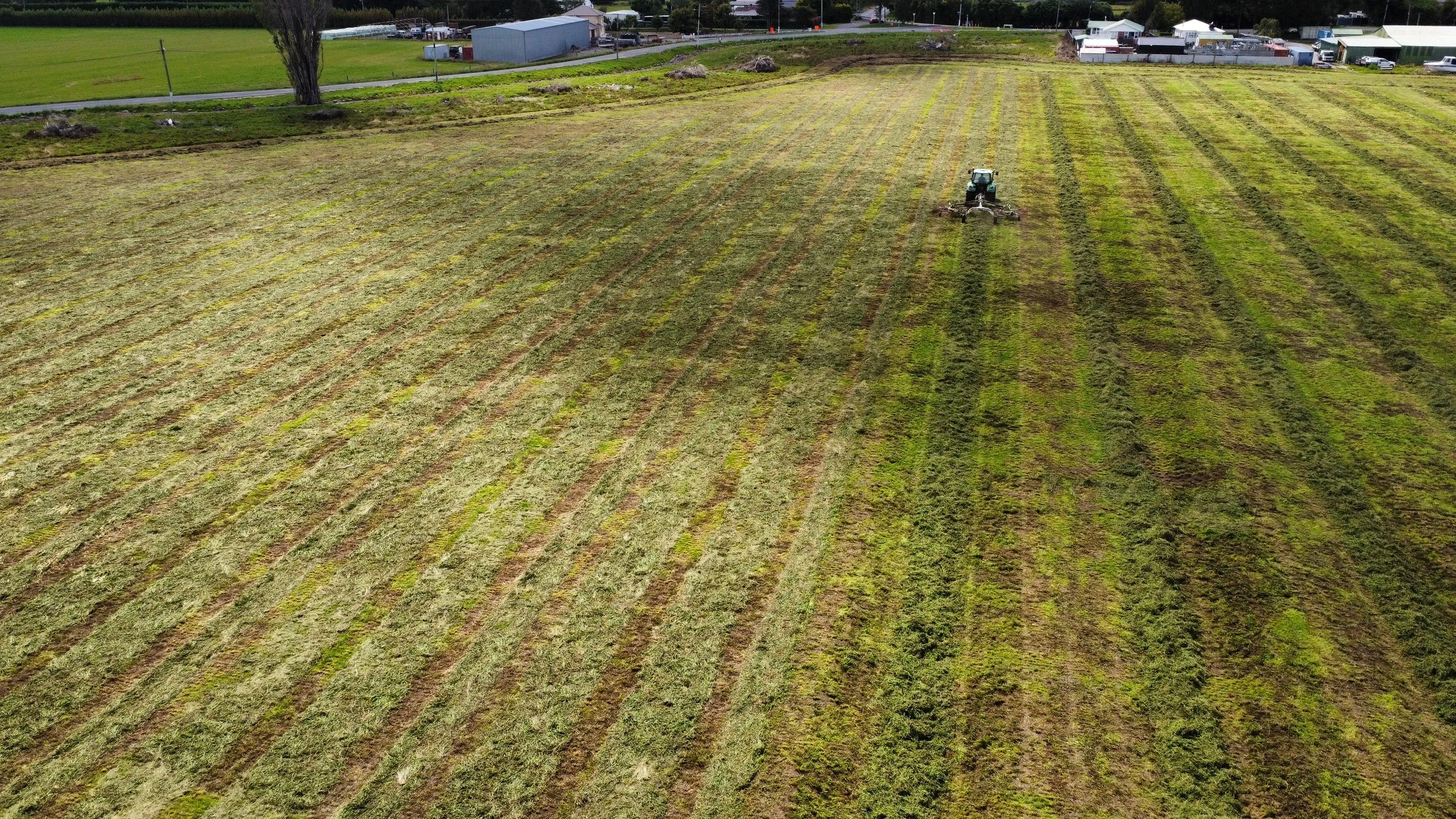 An aerial view of a large farm field being cultivated by a tractor that is tilling the soil, with rows of freshly disturbed earth. In the background, there are buildings, trees, and a road.