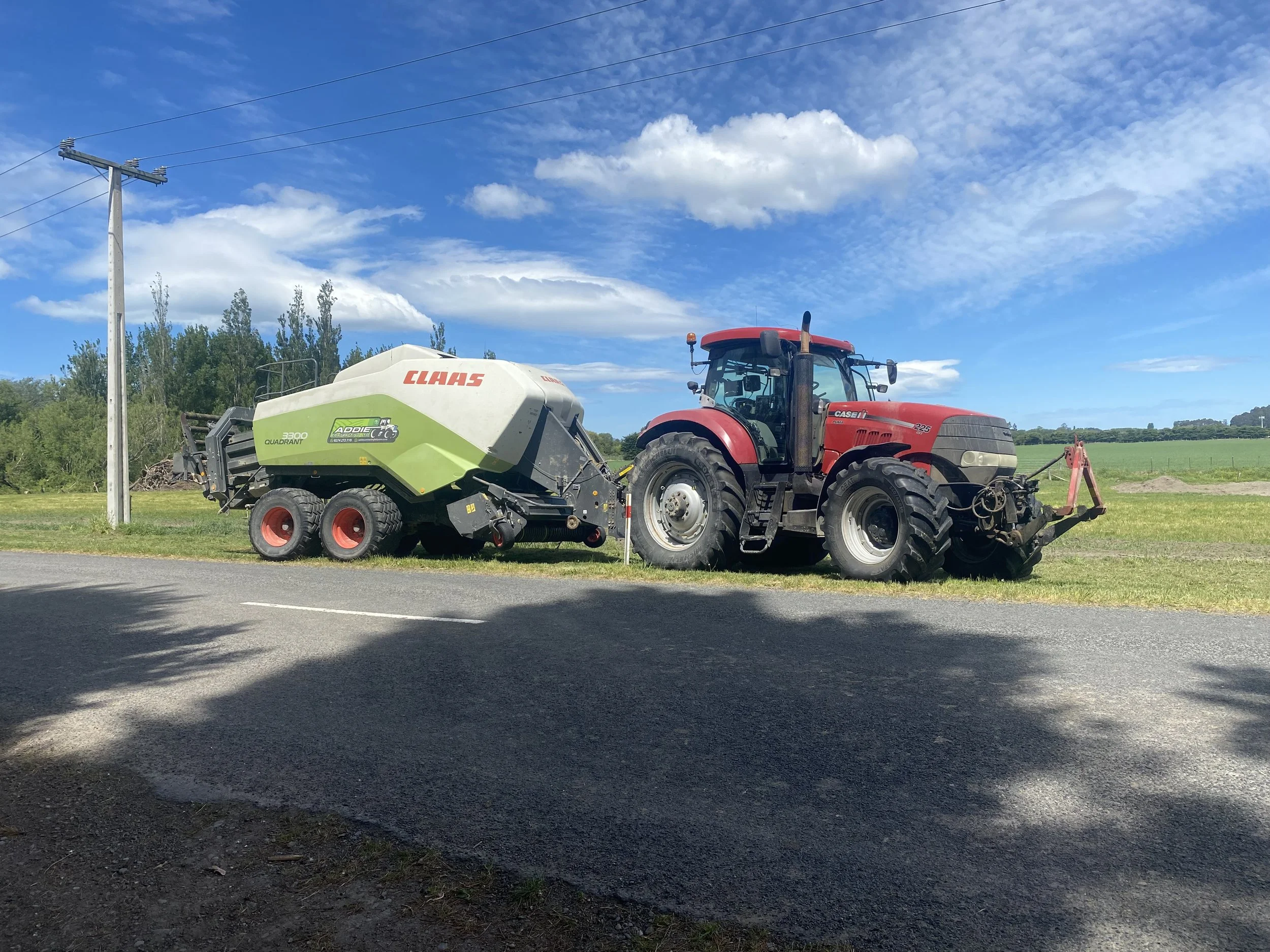 Red tractor parked on grass beside a green and white CLAAS baler on a rural road, with trees and open fields in the background under a partly cloudy sky.