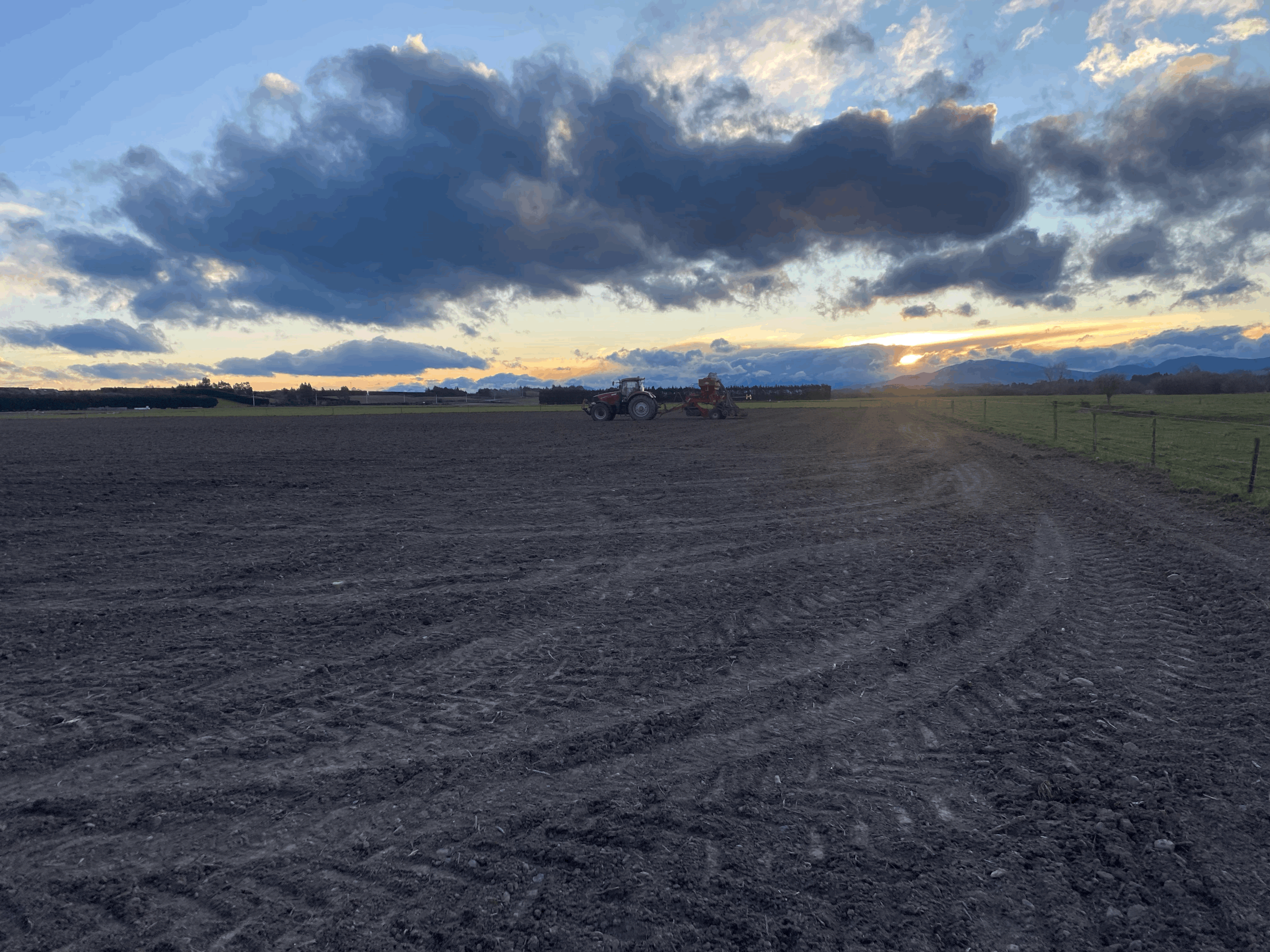 Farming field at sunset with a tractor and machinery in the background, expansive sky with dark clouds and some sunlight breaking through.