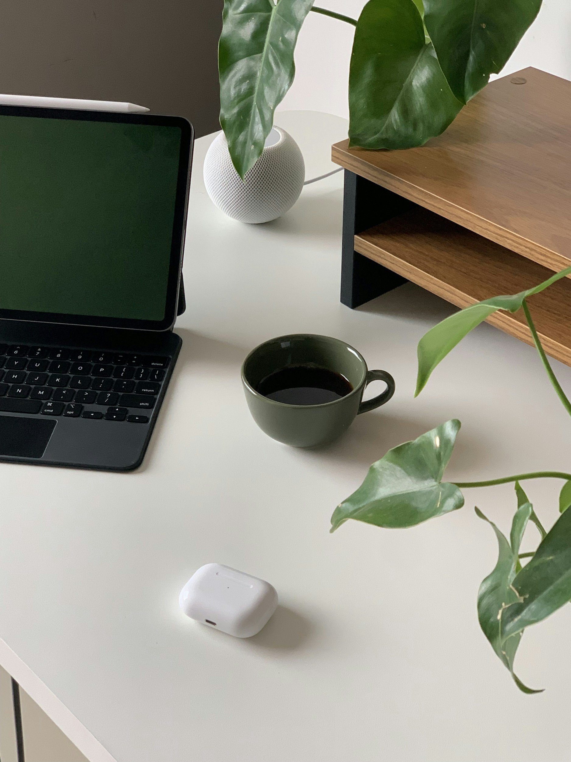 A minimalist workspace with a laptop, a cup of coffee, a white wireless earbud case, a potted plant, and a wooden shelf on a white desk.