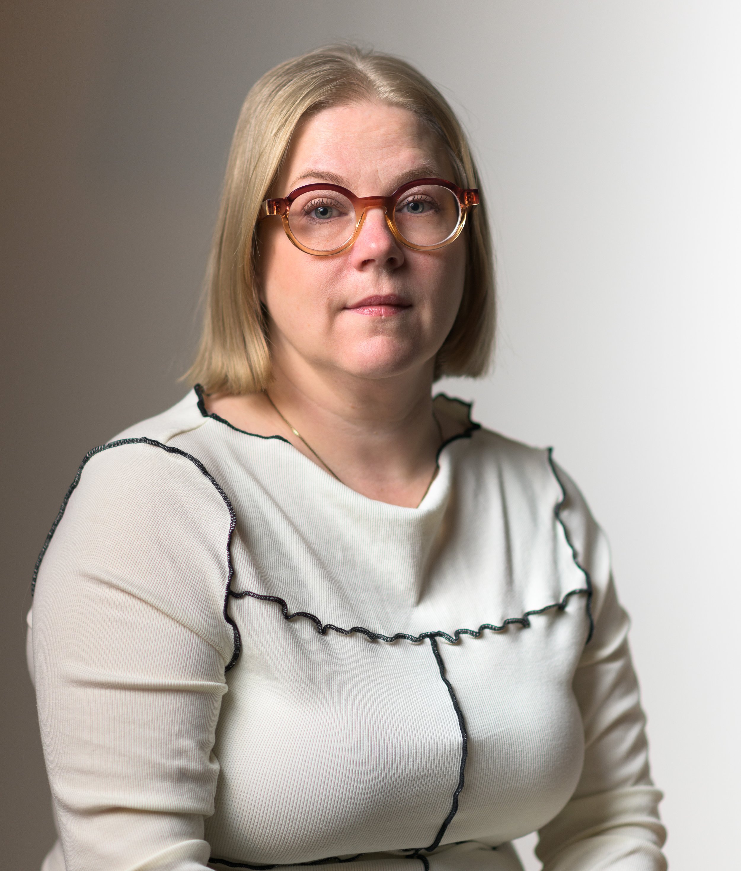 A woman with shoulder-length blonde hair and glasses, wearing a cream-colored top with black trim, sits against a light background.